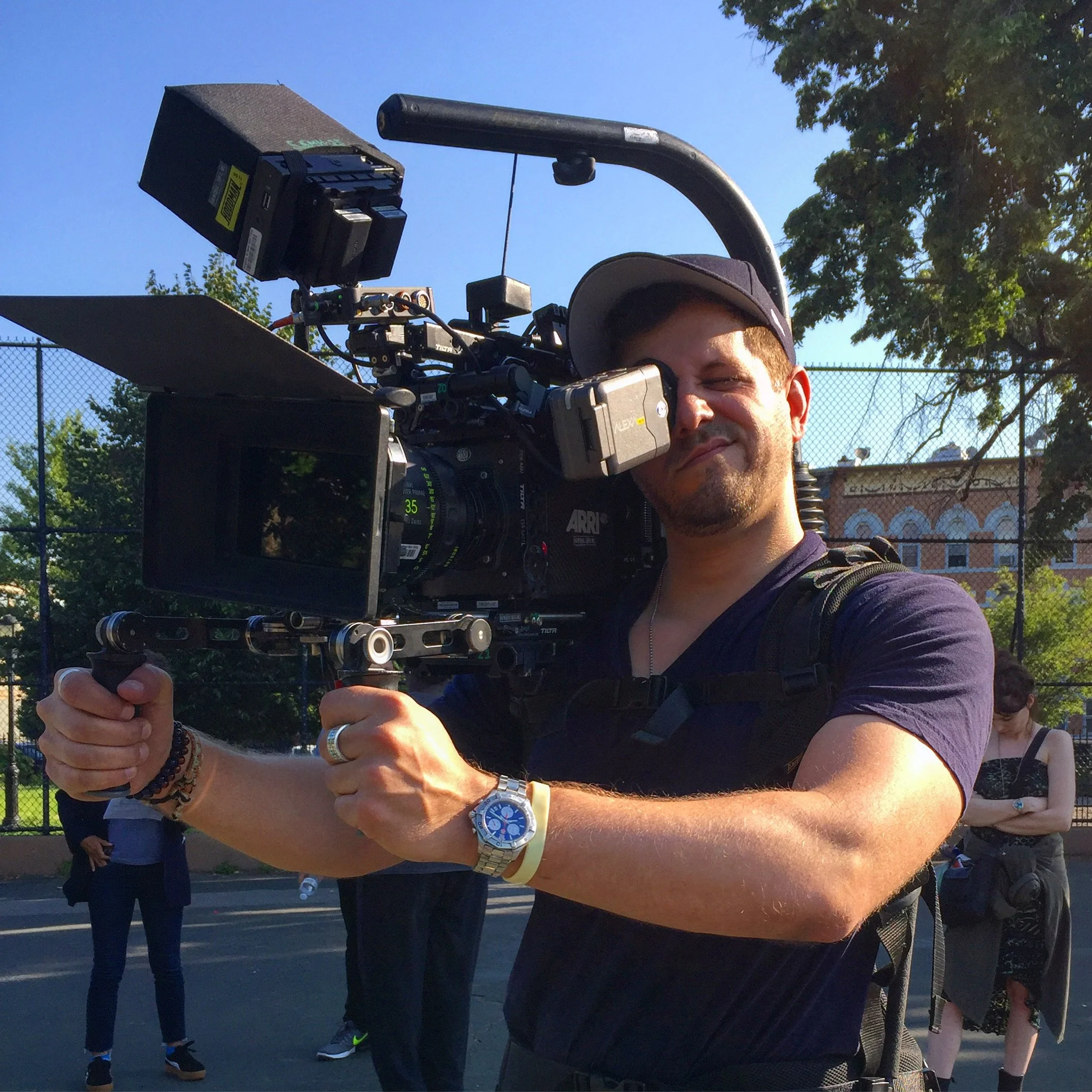 Man operating a professional video camera outdoors on a sunny day, with people in the background and trees.