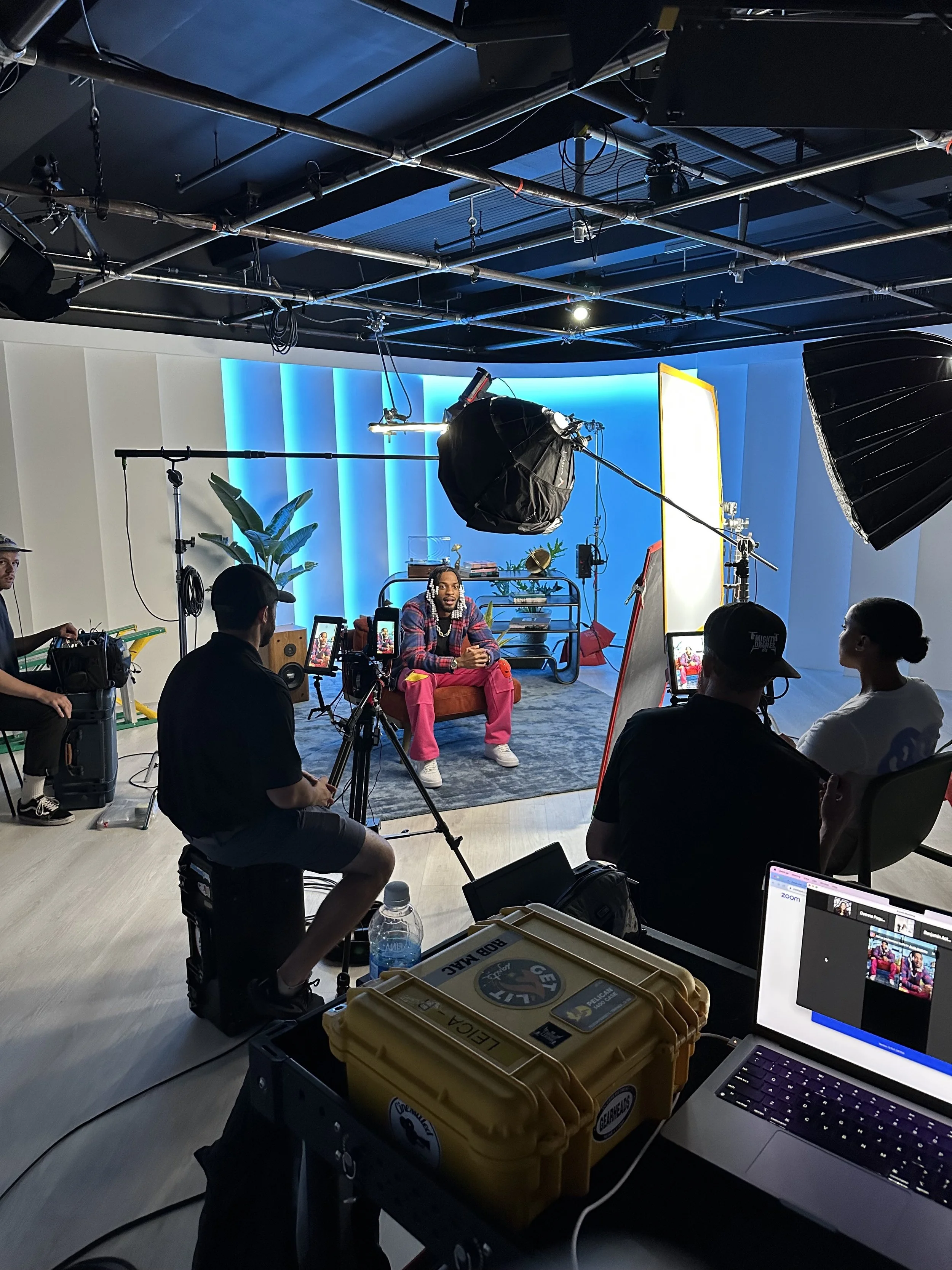 A person being filmed in a studio with technical crew, lighting, and camera equipment, seated on a red sofa with a blue illuminated backdrop.