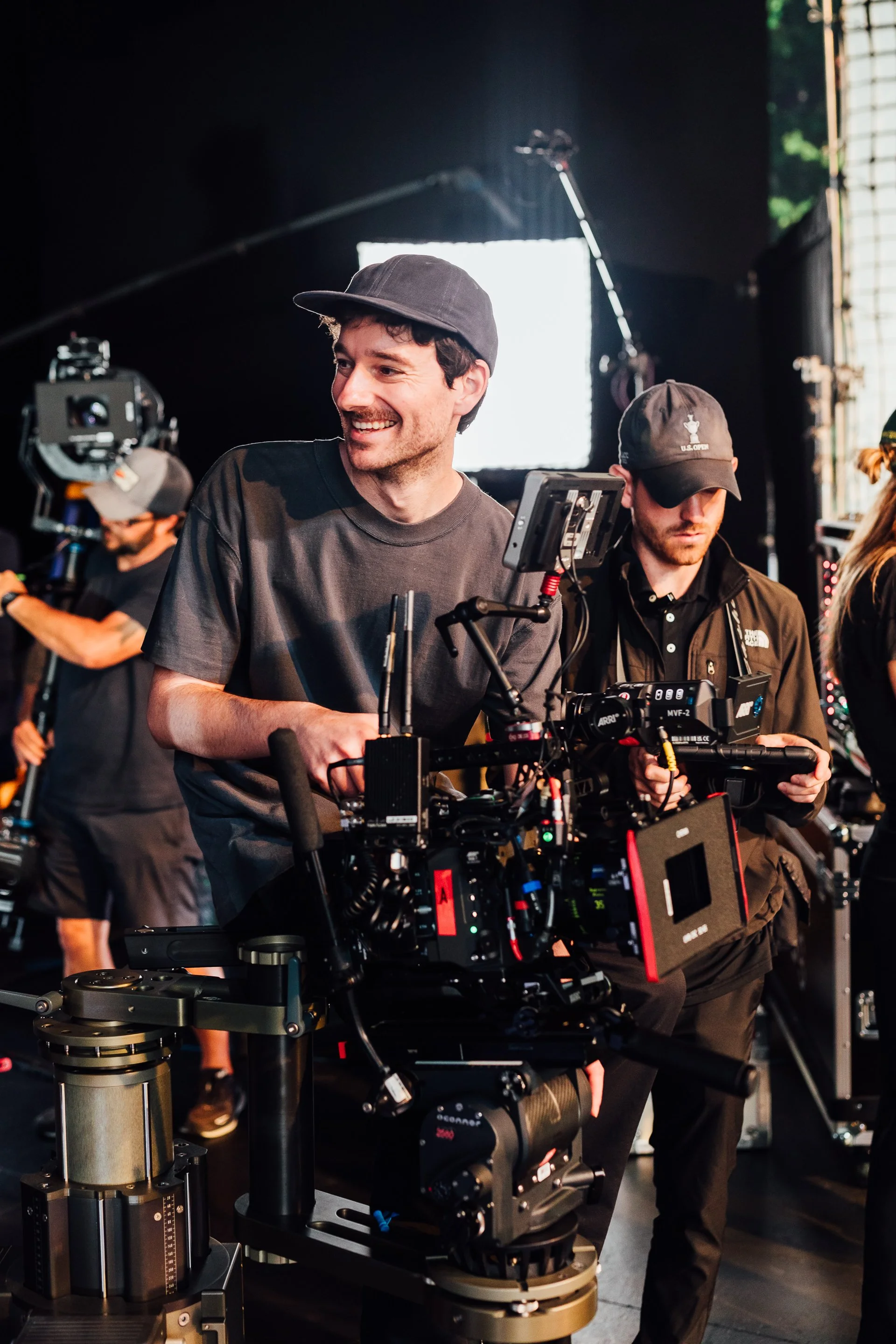Film crew working on set with film equipment, cameras, and monitors in studio, crew members wearing black caps and casual clothing, one smiling and looking at camera.