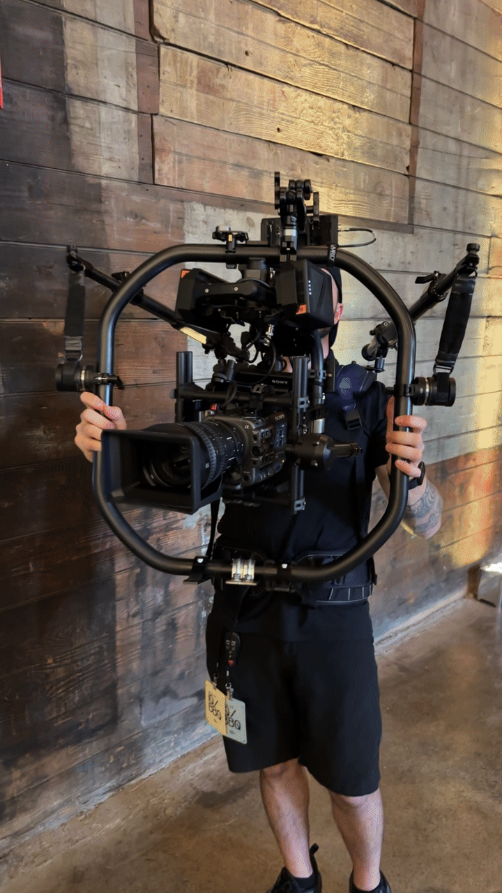A person holding a large camera stabilizer rig with a professional video camera attached, standing in front of a wooden wall.