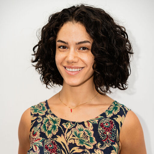 A woman smiling with messy dark hair, wearing a plaid jacket over a white shirt, against a textured blue background.
