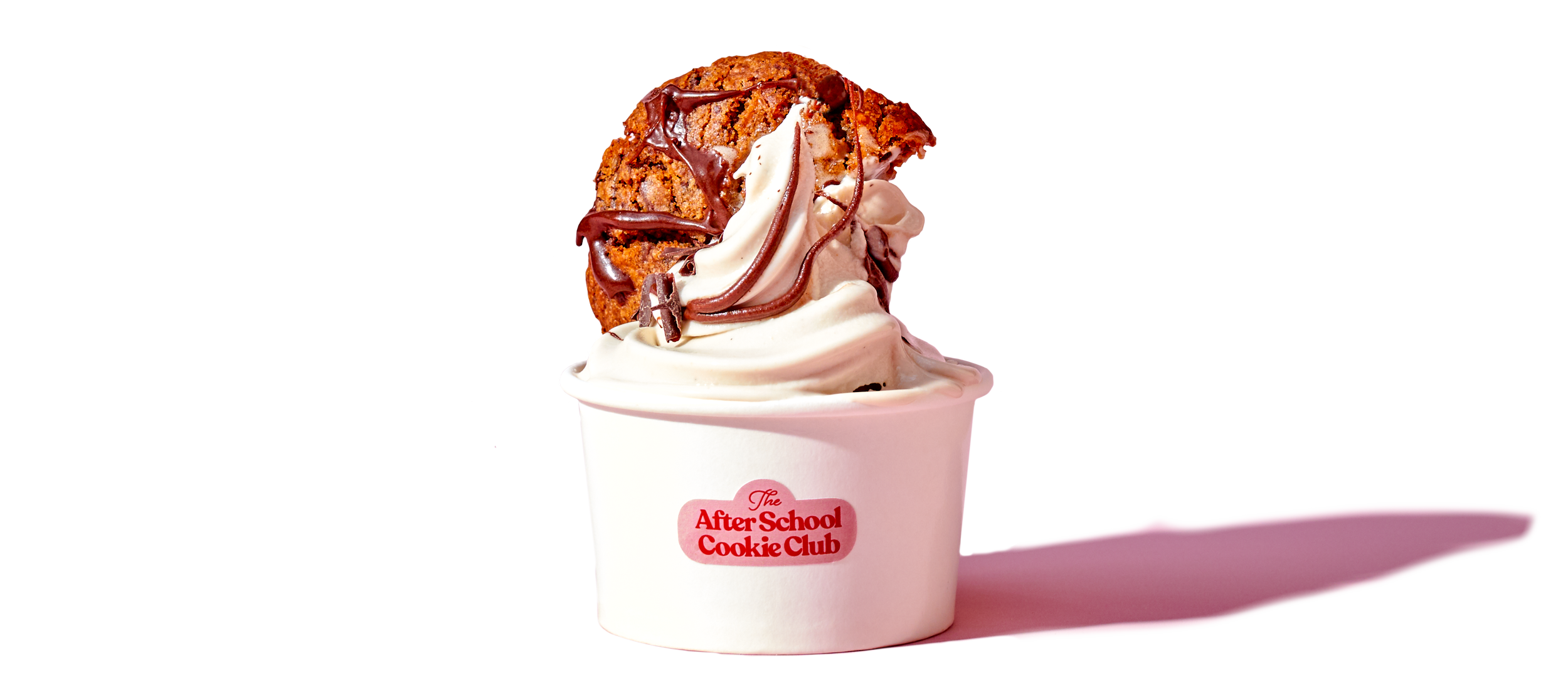 Ice cream cup with chocolate chip cookie on top, drizzled with chocolate and caramel, in a white cup labeled "The After School Cookie Club" on a pink background.