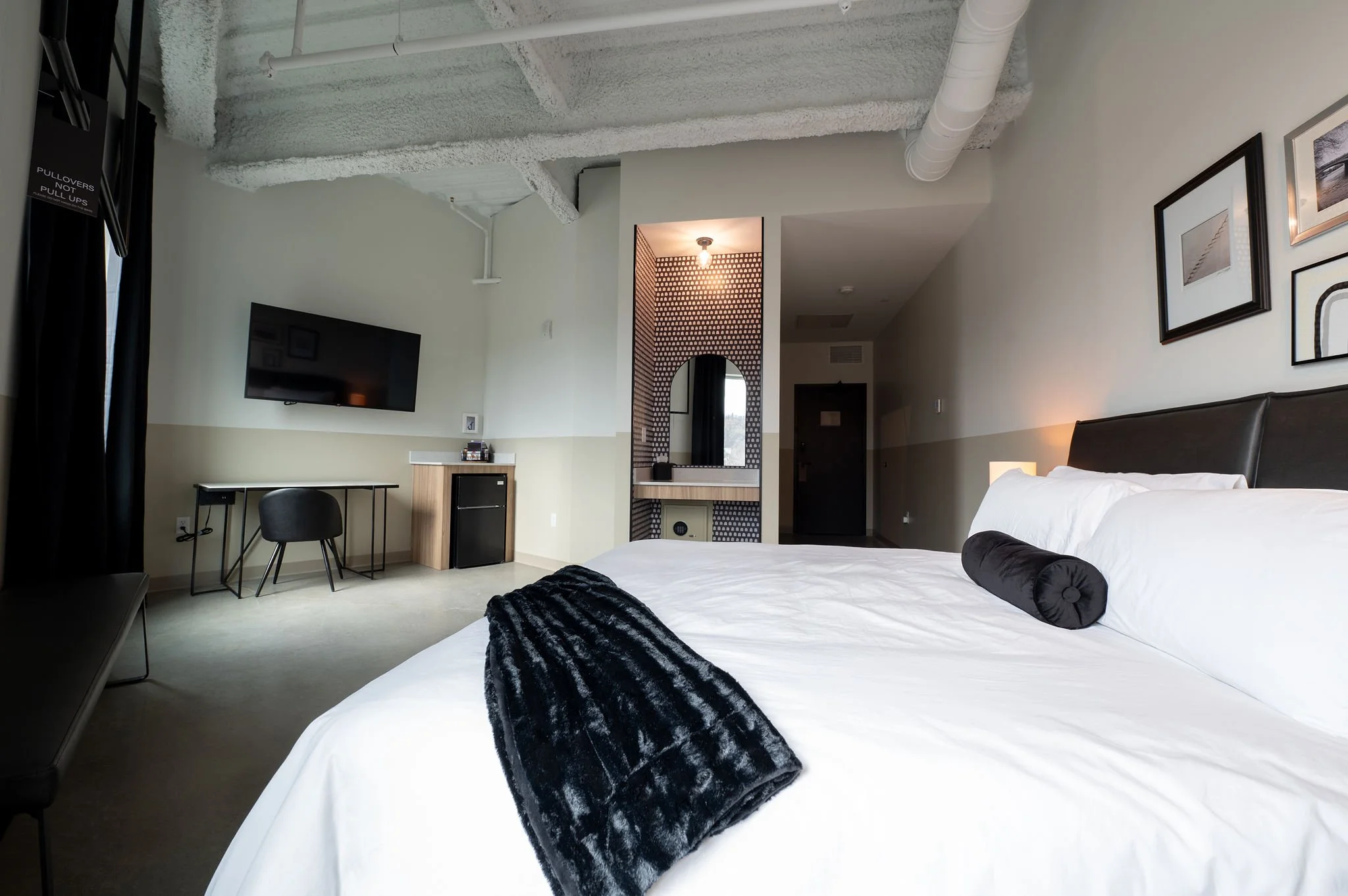 Modern hotel room with a large white bed, black bolster pillow, black throw blanket, wall-mounted TV, small desk with a chair, and decorative wall art.