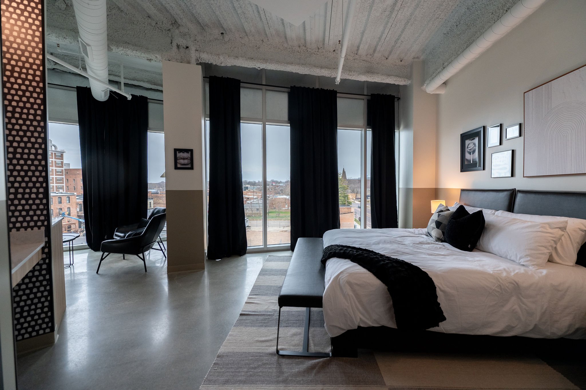 Modern hotel room with a large bed, black and white pillows, a small bench at the foot of the bed, and a nightstand with a lamp. Floor-to-ceiling windows with black curtains, revealing an outdoor city view. Artworks on the wall and exposed ceiling pipes.