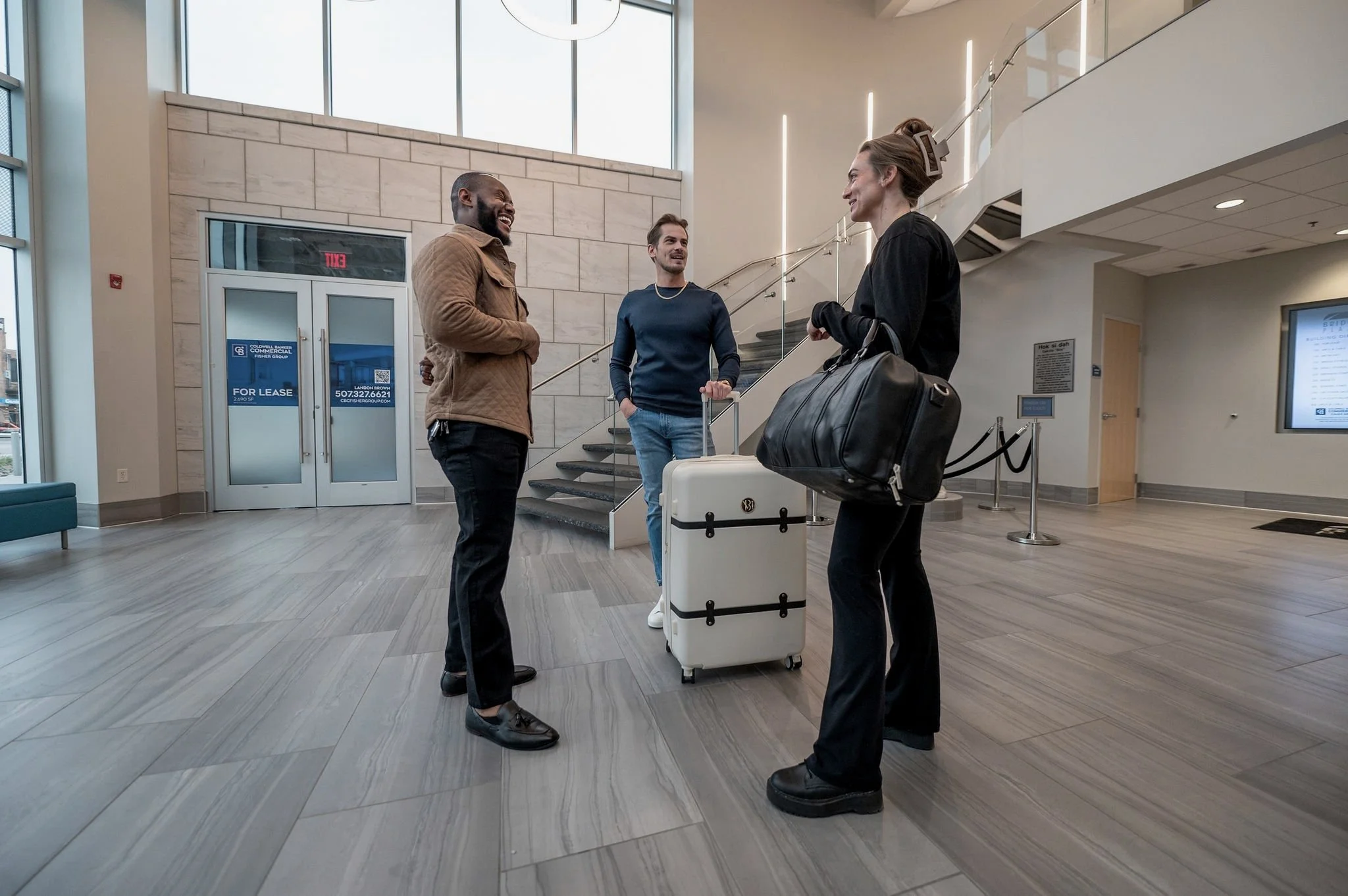 Three people with luggage chatting inside a modern building's lobby.