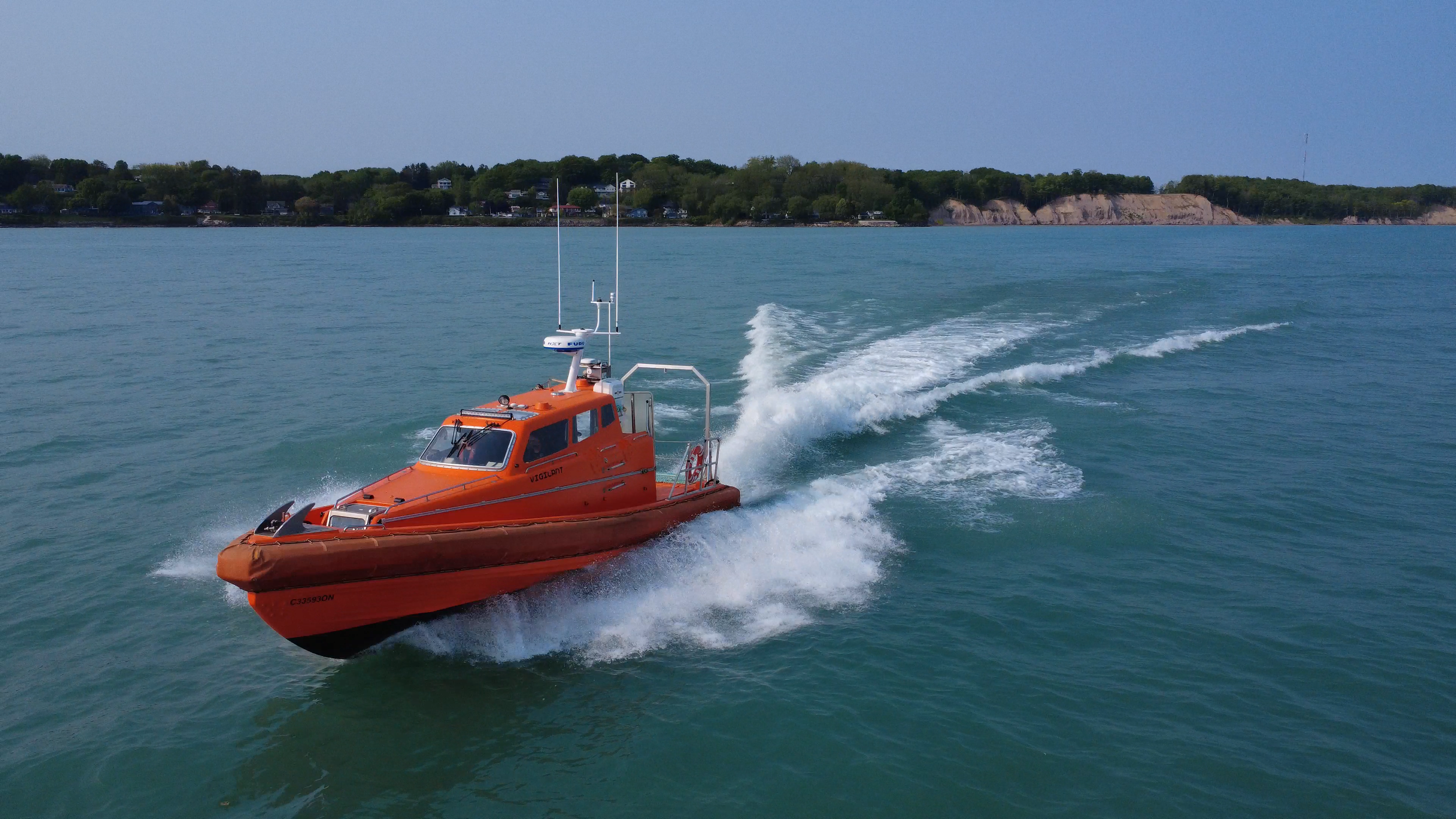 An orange rescue boat speeding across the water with a wake behind it, near a shoreline with trees and houses.