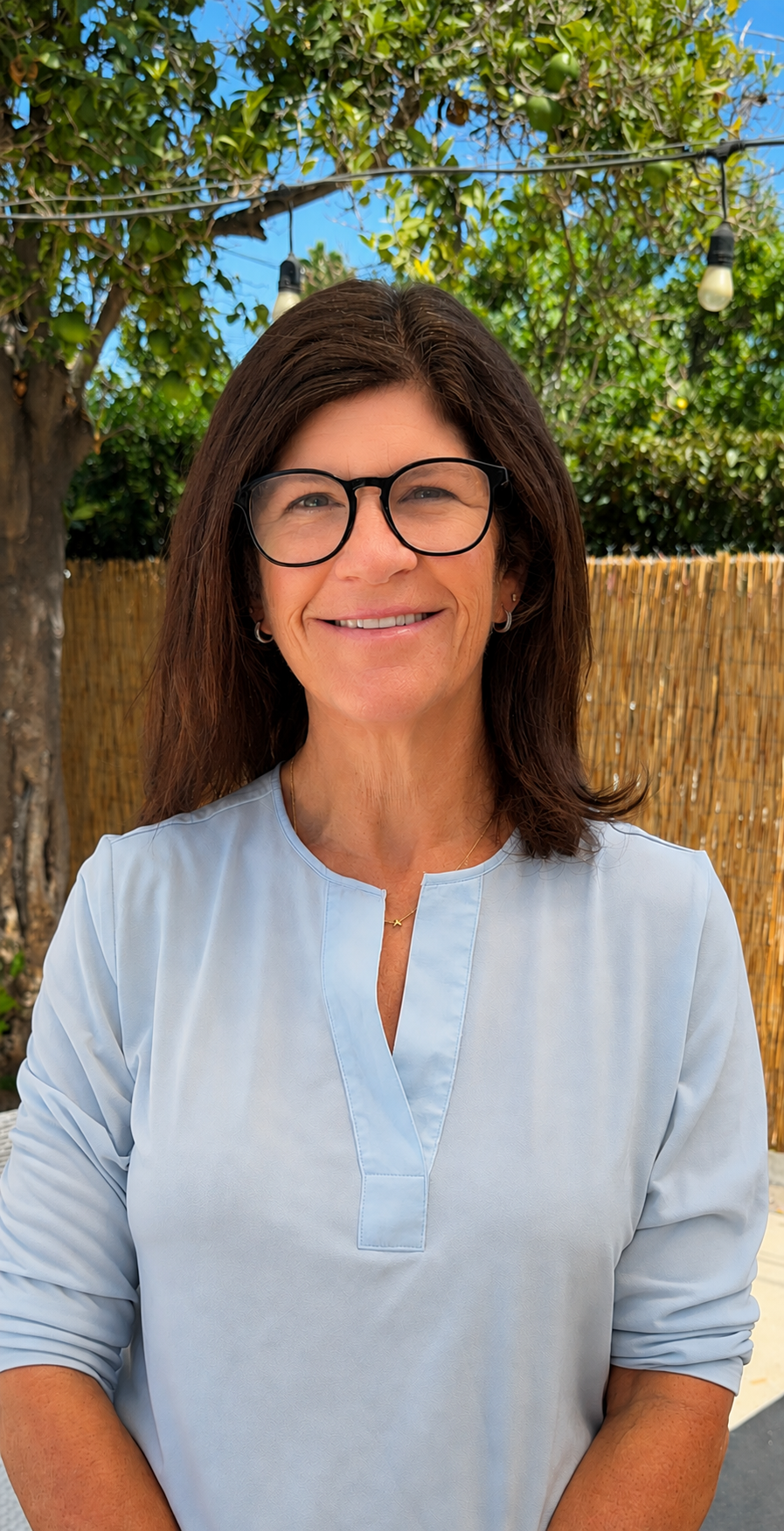 A woman with brown hair, glasses, and wearing a light blue blouse smiling outdoors with trees and string lights in the background.