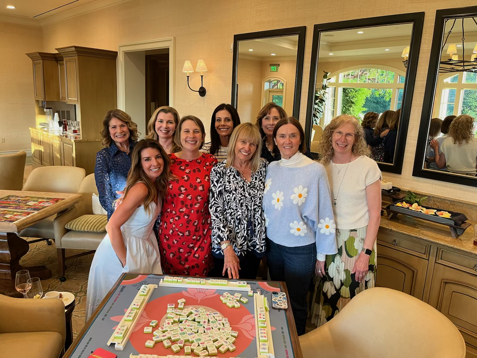 Group of women gathered in a restaurant or banquet room, smiling for a photo, with a Mahjong game setup on the table in front of them.