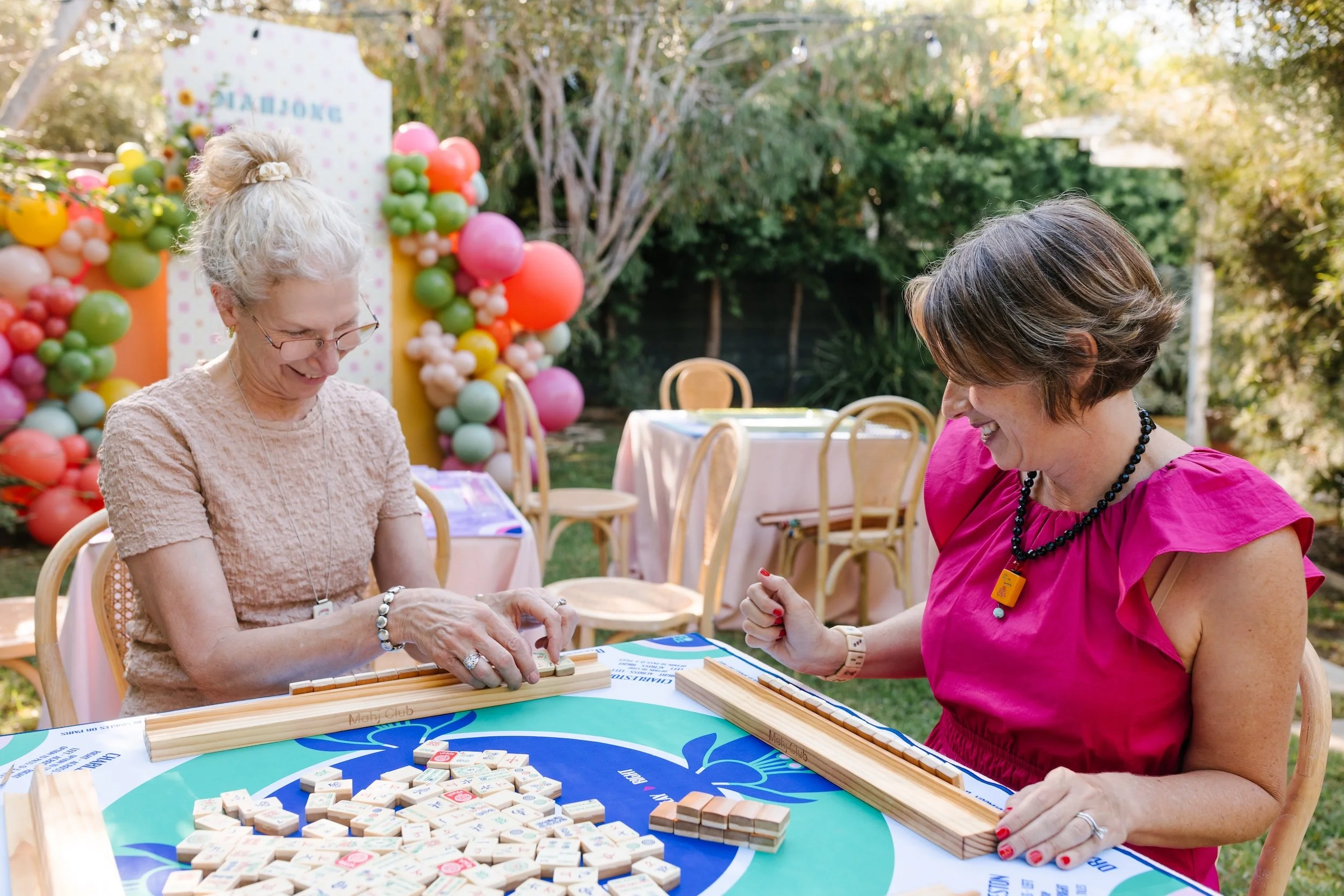 Mahj Club Founder Sara Linden and Instructor Carol Weinfeld playing American Mah Jongg together at a colorful outdoor event in Orange County, California, surrounded by balloons and mahjong tiles — promoting community through play.