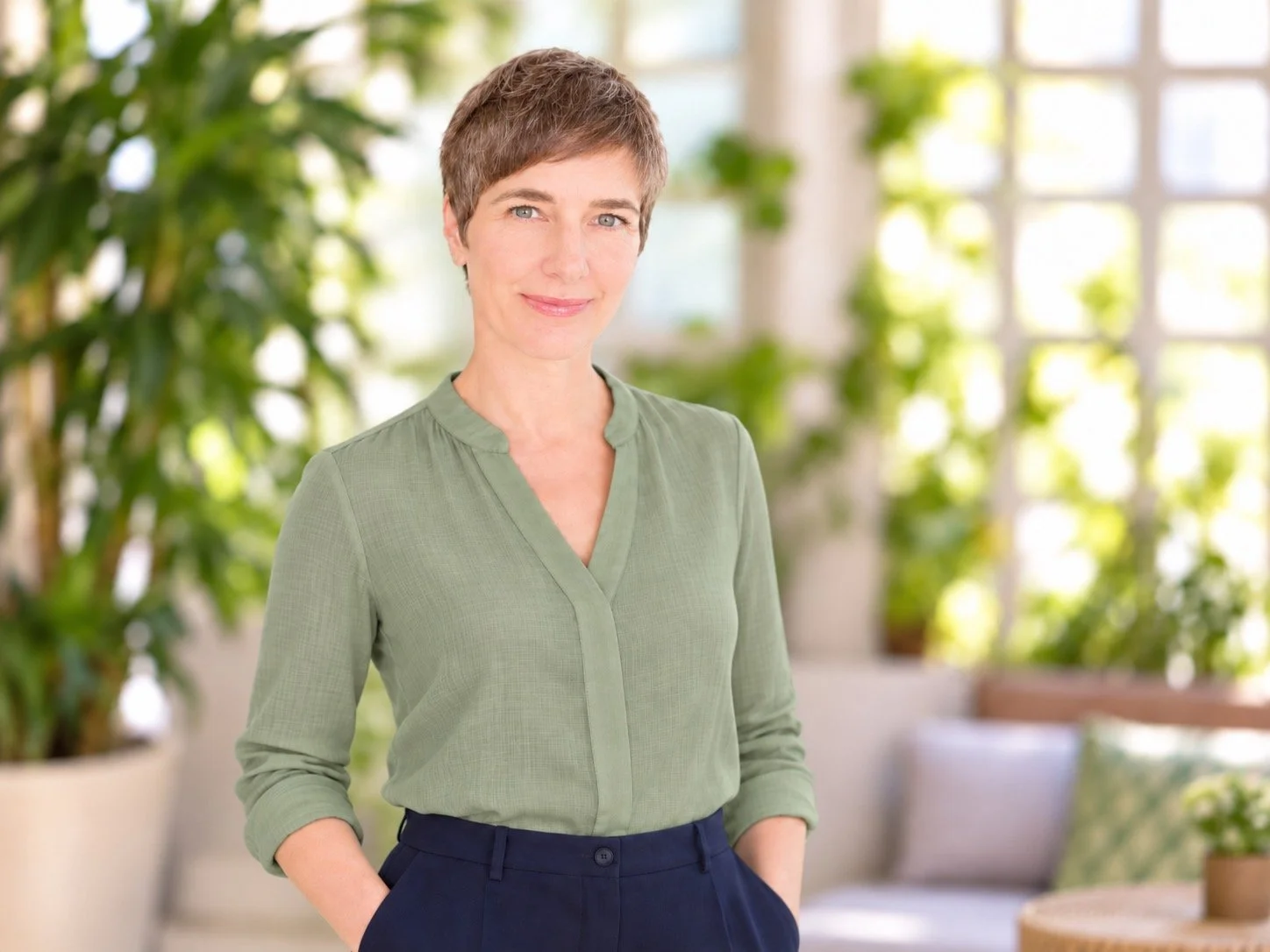 A woman with short brown hair and blue eyes standing indoors in front of green plants, wearing a light green blouse and dark pants, with a soft smile.