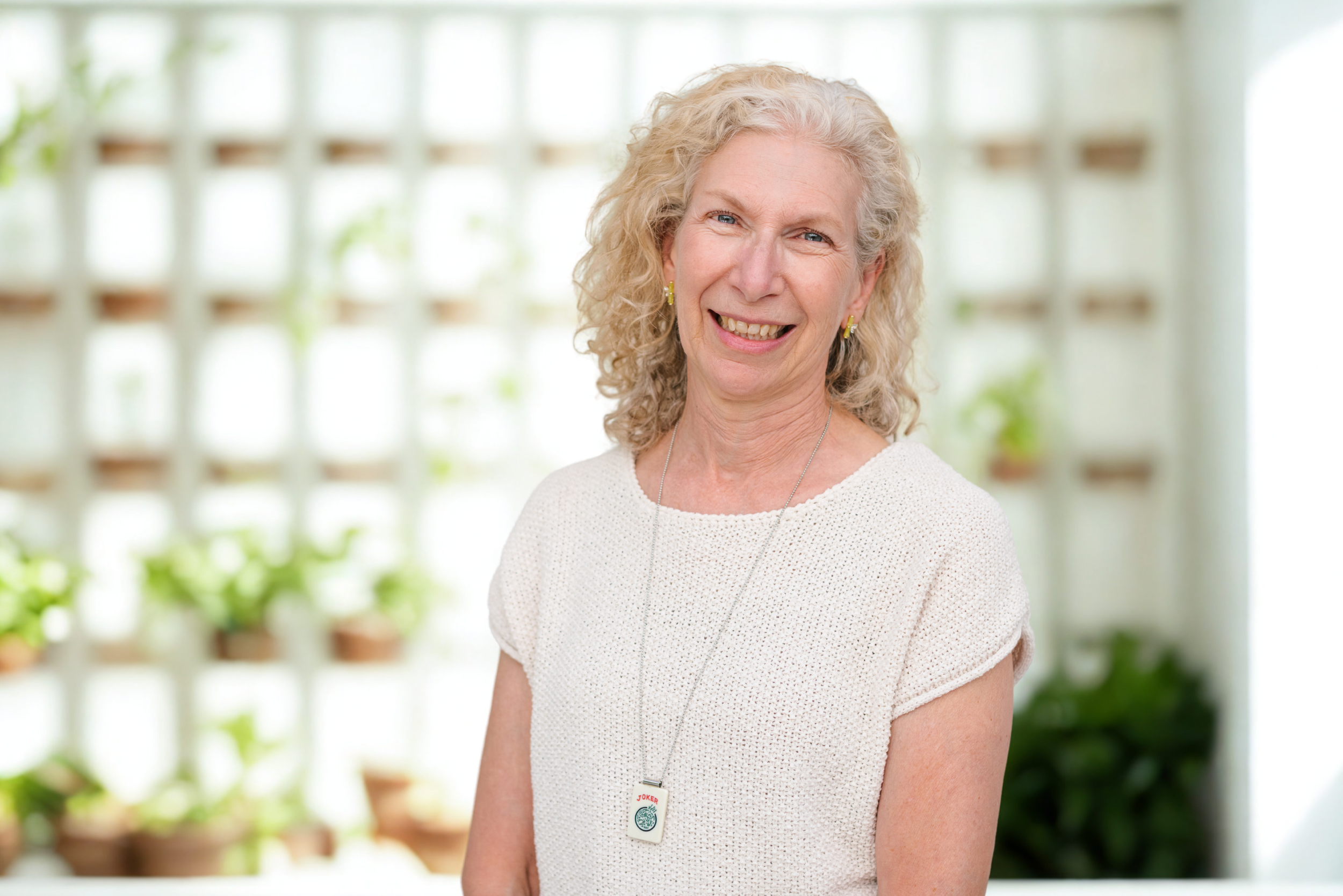 Carol Weinfeld, Mahj Club Instructor, smiling in a soft-lit studio portrait wearing a cream knit top and mahjong “joker” tile necklace — American Mah Jongg instructor in Southern California