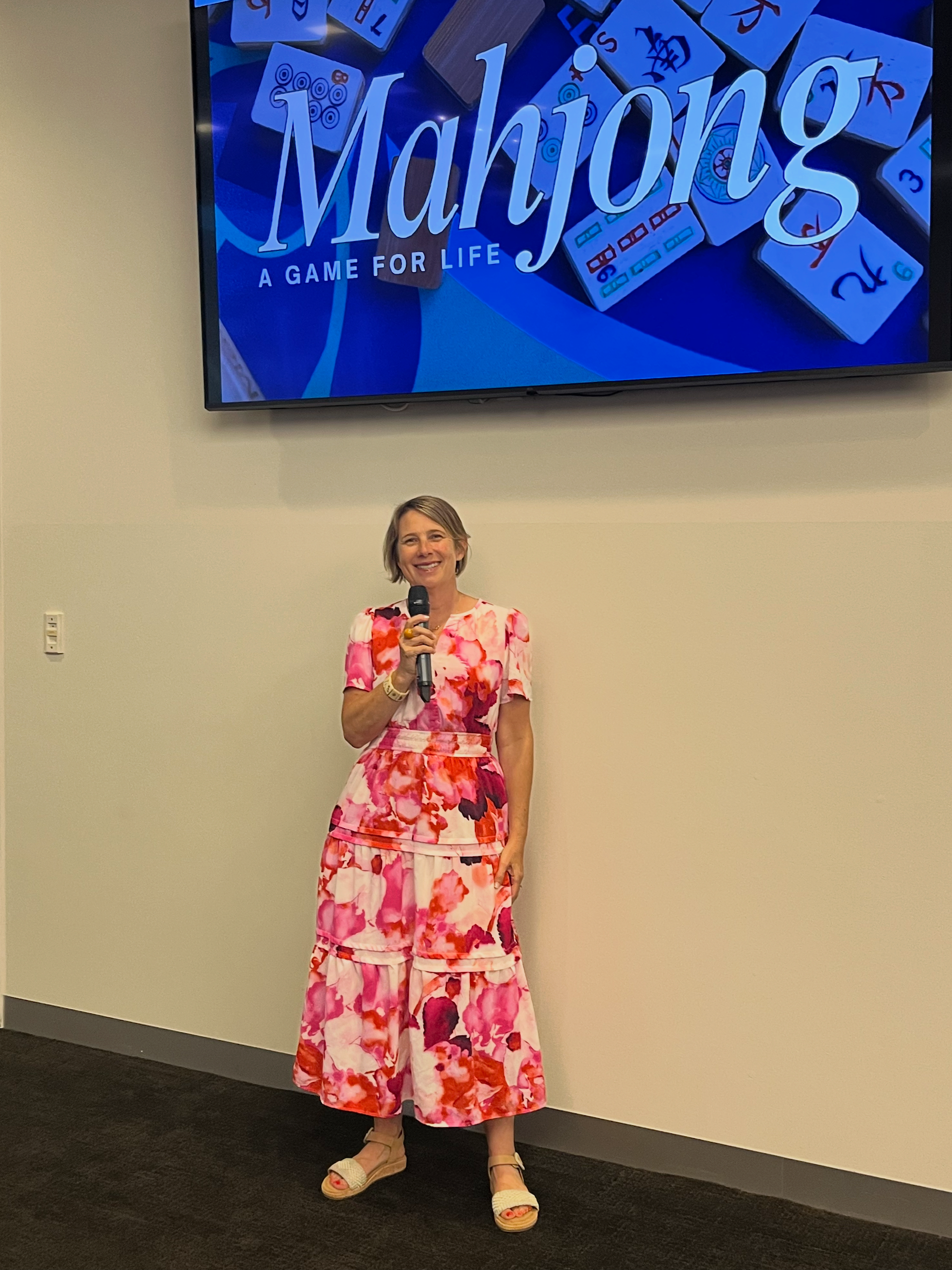 A woman in a colorful pink, red, and white floral dress is standing in front of a wall, holding a microphone and smiling. A large screen behind her displays the title 'Mahjong: A Game for Life' with images of mahjong tiles.