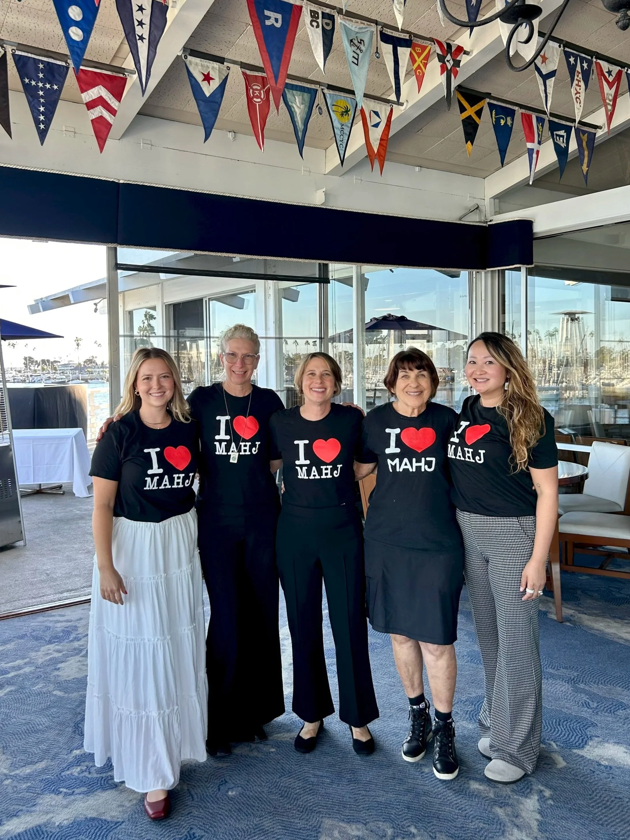 Five women standing together at a corporate mahjong event, wearing matching black T-shirts that say 'I ❤️ MAHJ'. Behind them are large windows showing a marina with boats and palm trees, and colorful pennant banners hanging from the ceiling.