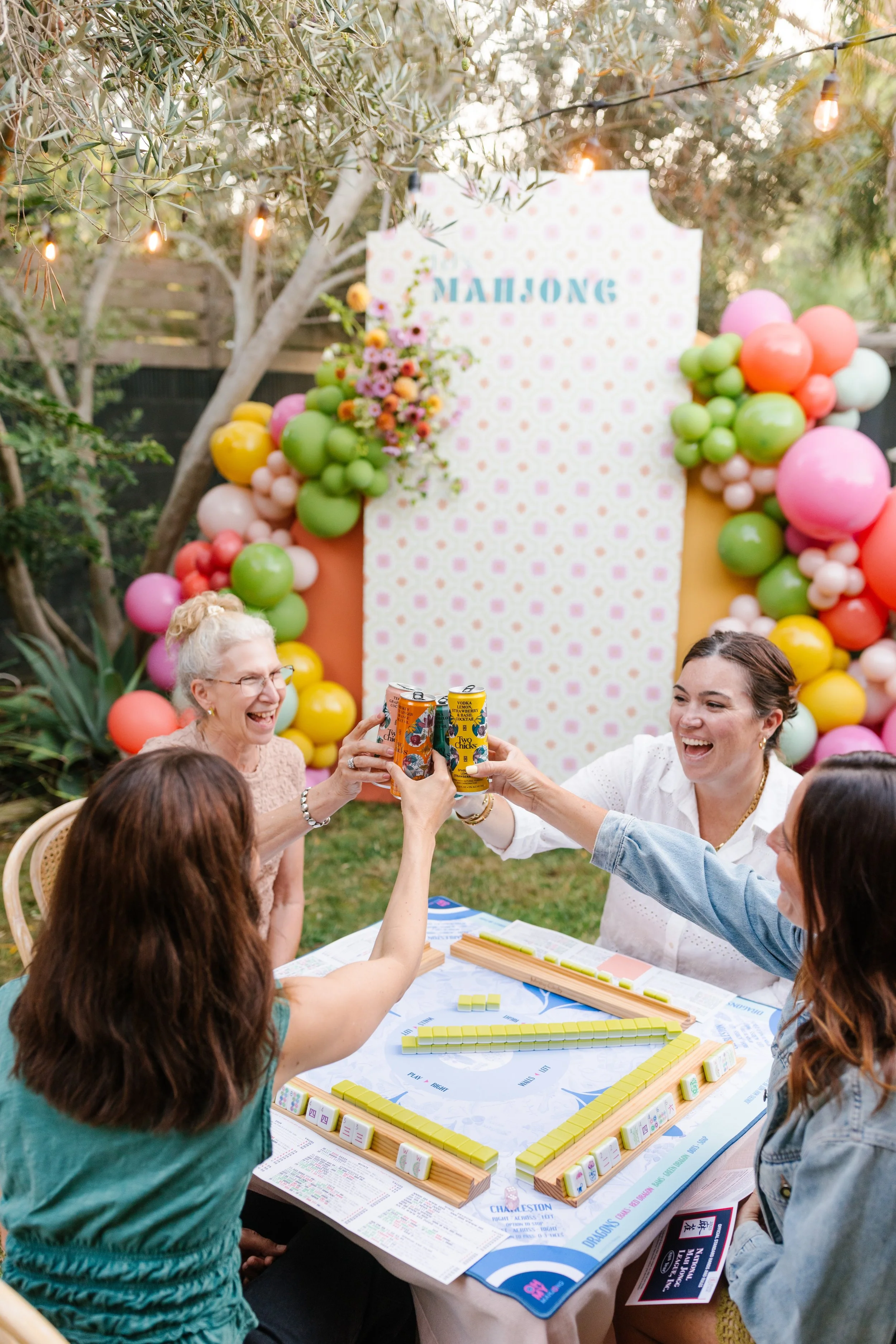 People gathered outdoors at a table playing a game of Mahjong, with balloons and a floral backdrop, to celebrate a special occasion.