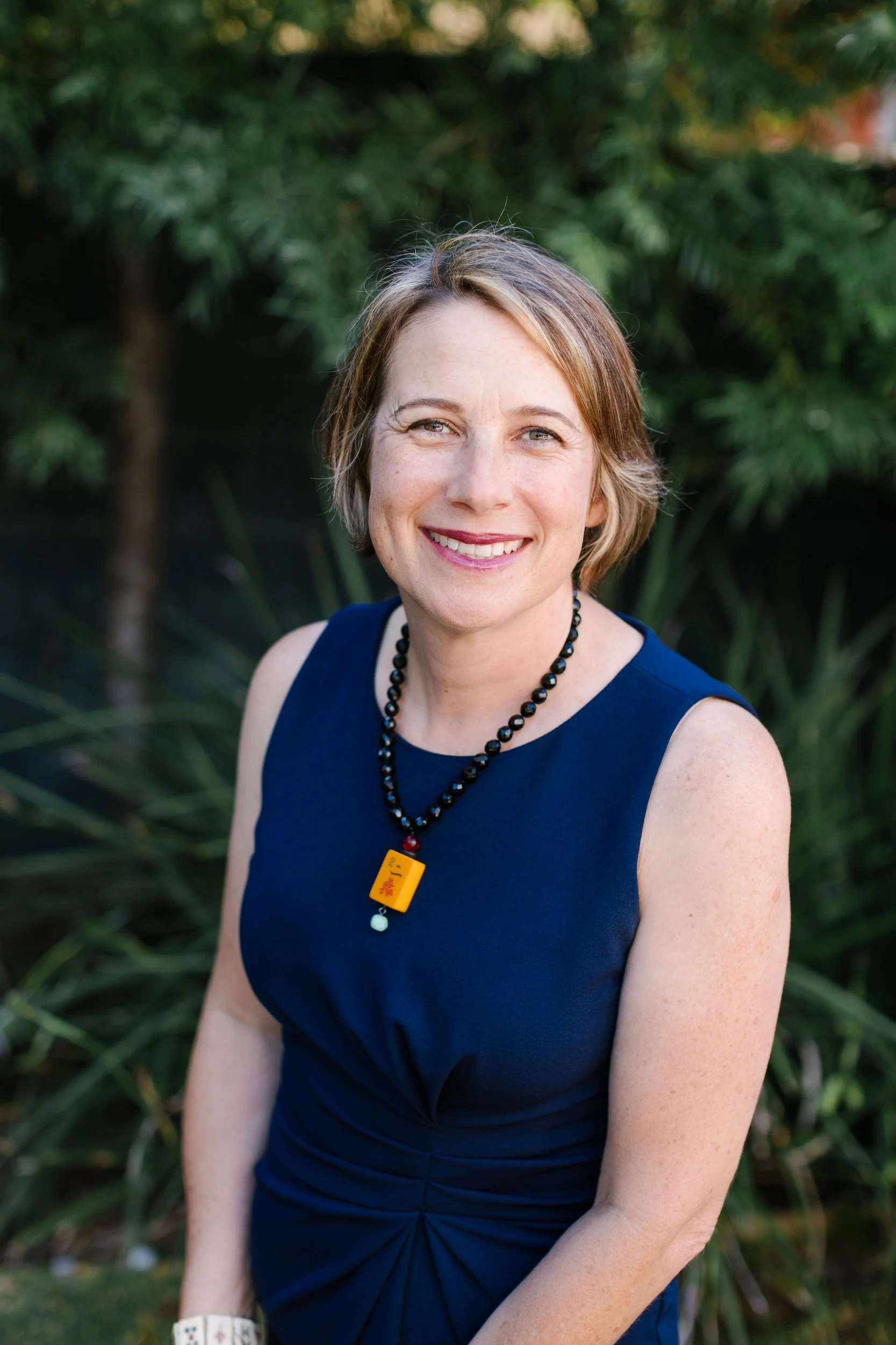 Sara Linden, Founder and Instructor at Mahj Club, smiling outdoors in navy dress and mahjong tile necklace — teaching and promoting American Mah Jongg community in Southern California.