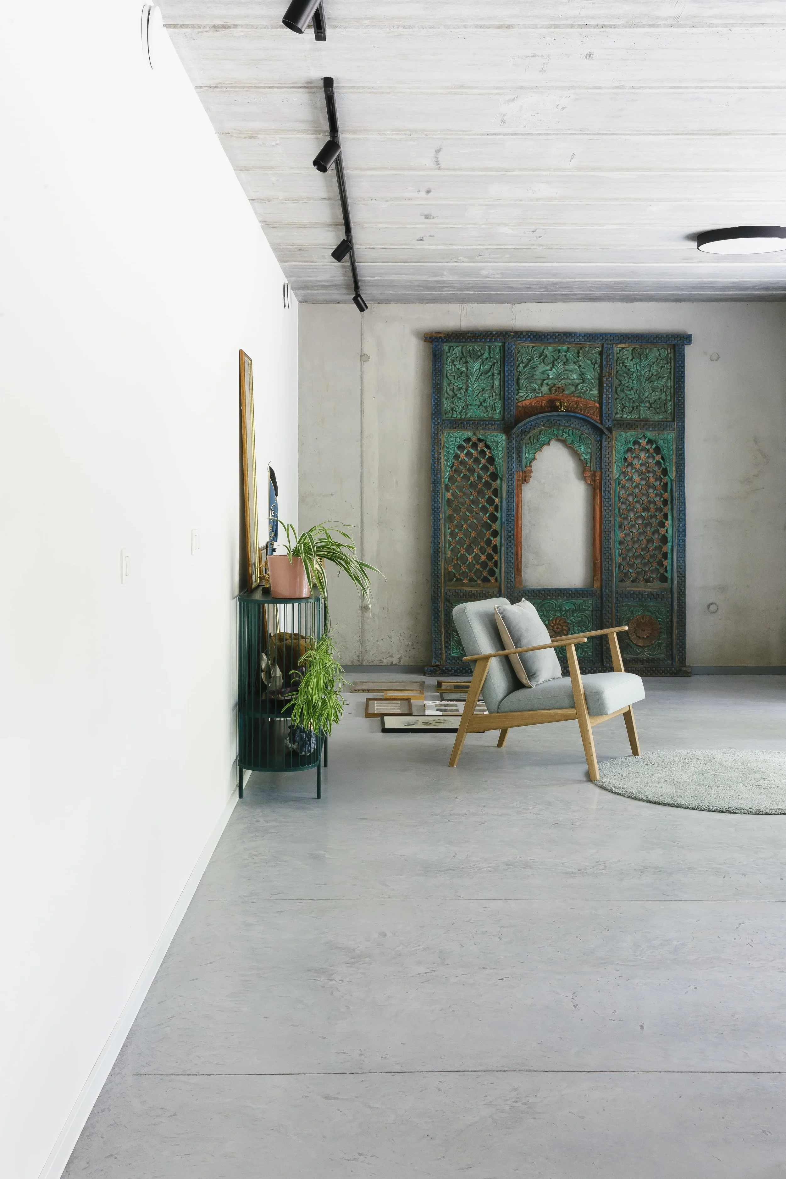 Een binnentuin met een lichte vloer en een witte muur. Er staat een groene plant op een bijzettafel, en er is een vintage decoratief houten scherm met ingelijste patronen. Een comfortabele armstoel met een kussen, en een ronde grijze tapijt op de vloer.