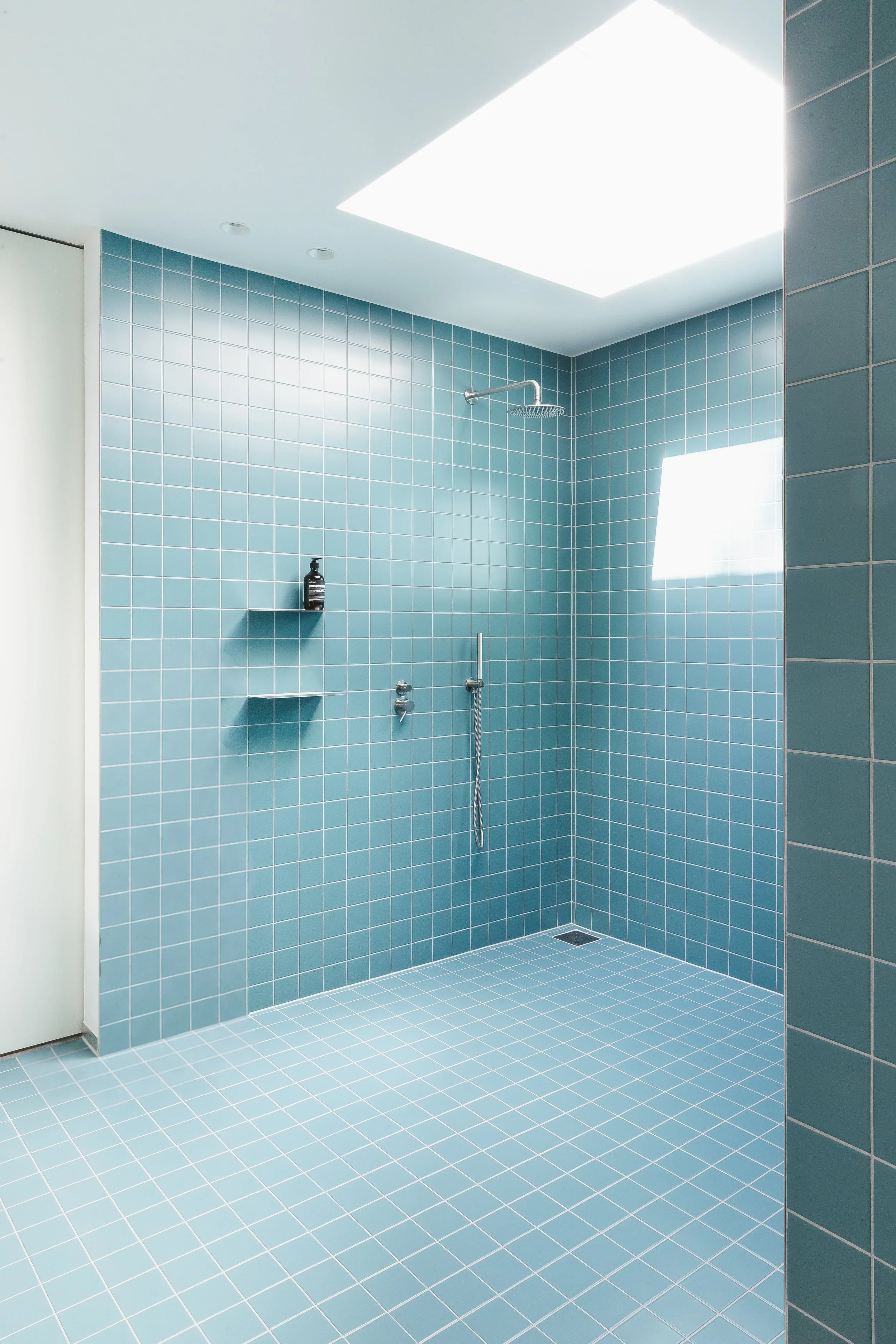 Een moderne badkamer met blauwe vierkante tegels op de muren en vloer, een inloopdouche met regen(douche)kop en een raam dat licht binnenlaat.