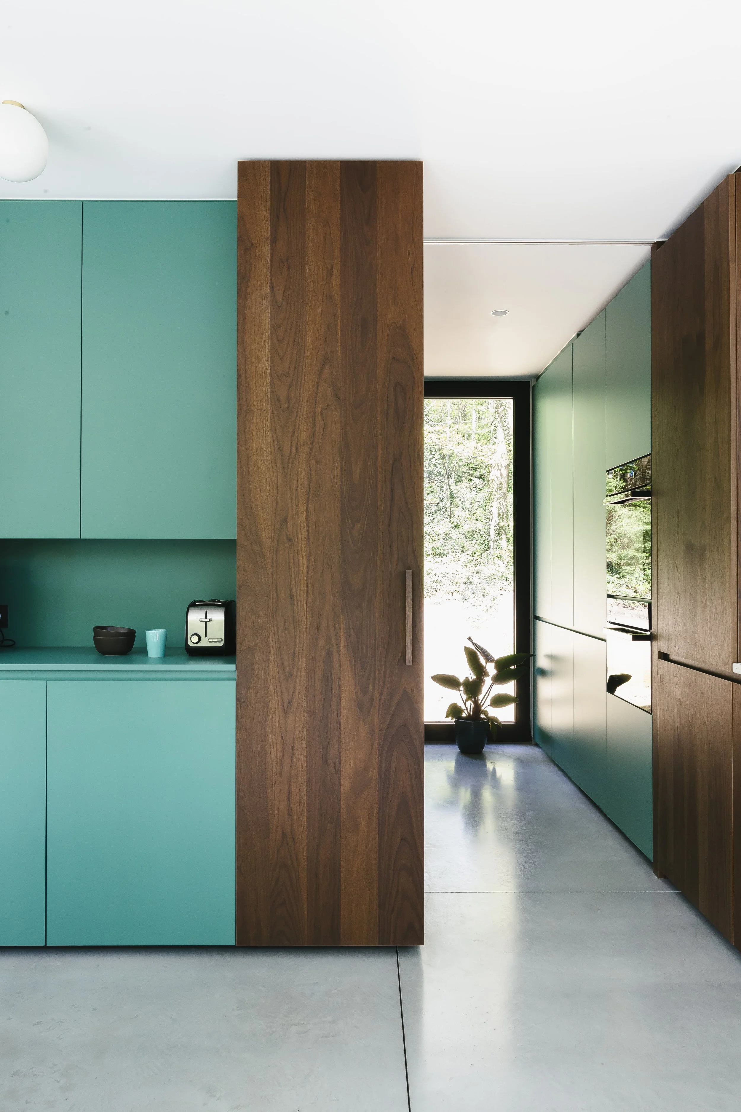 Interieur van een moderne keuken met groene kastdeuren, houten panelen en een grote raam met een uitzicht op groene bomen.