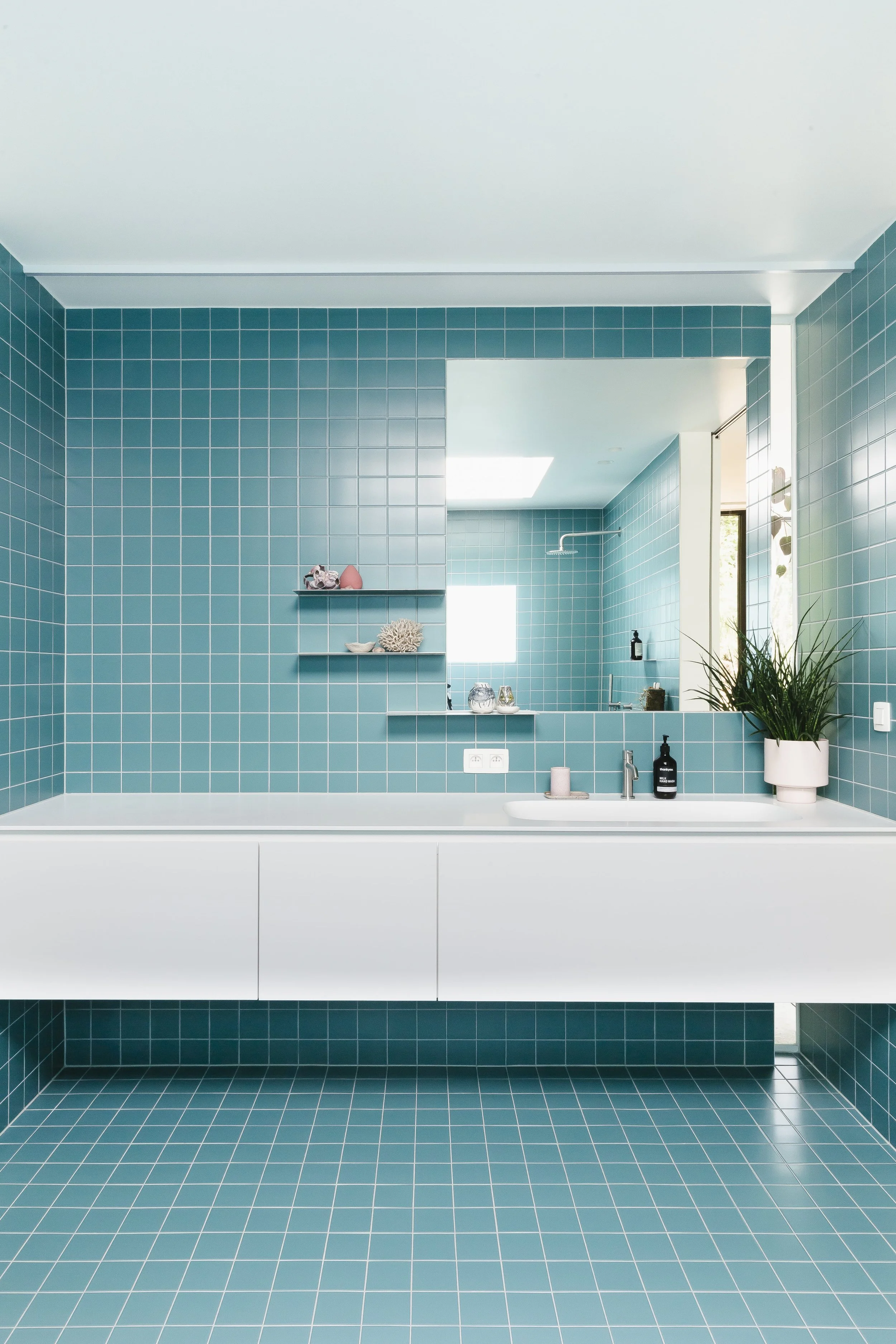 Een moderne badkamer met turquoise tegels op de muren en vloer, een bad met witte kast eronder, een spiegel, planten en decoraties.