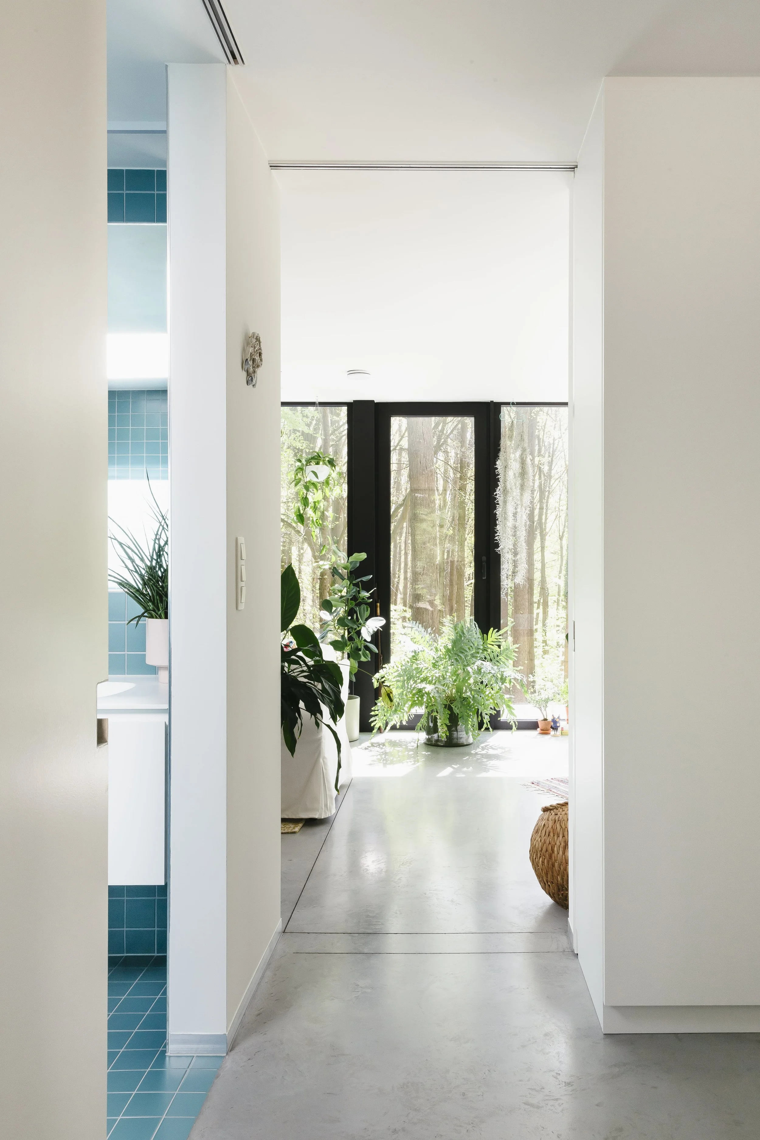 Binnenruimte met grote glazen deur die naar een tuin leidt, met diverse groene planten en een hoektapijt op de vloer, witte muren en een lichte, minimalistische inrichting.