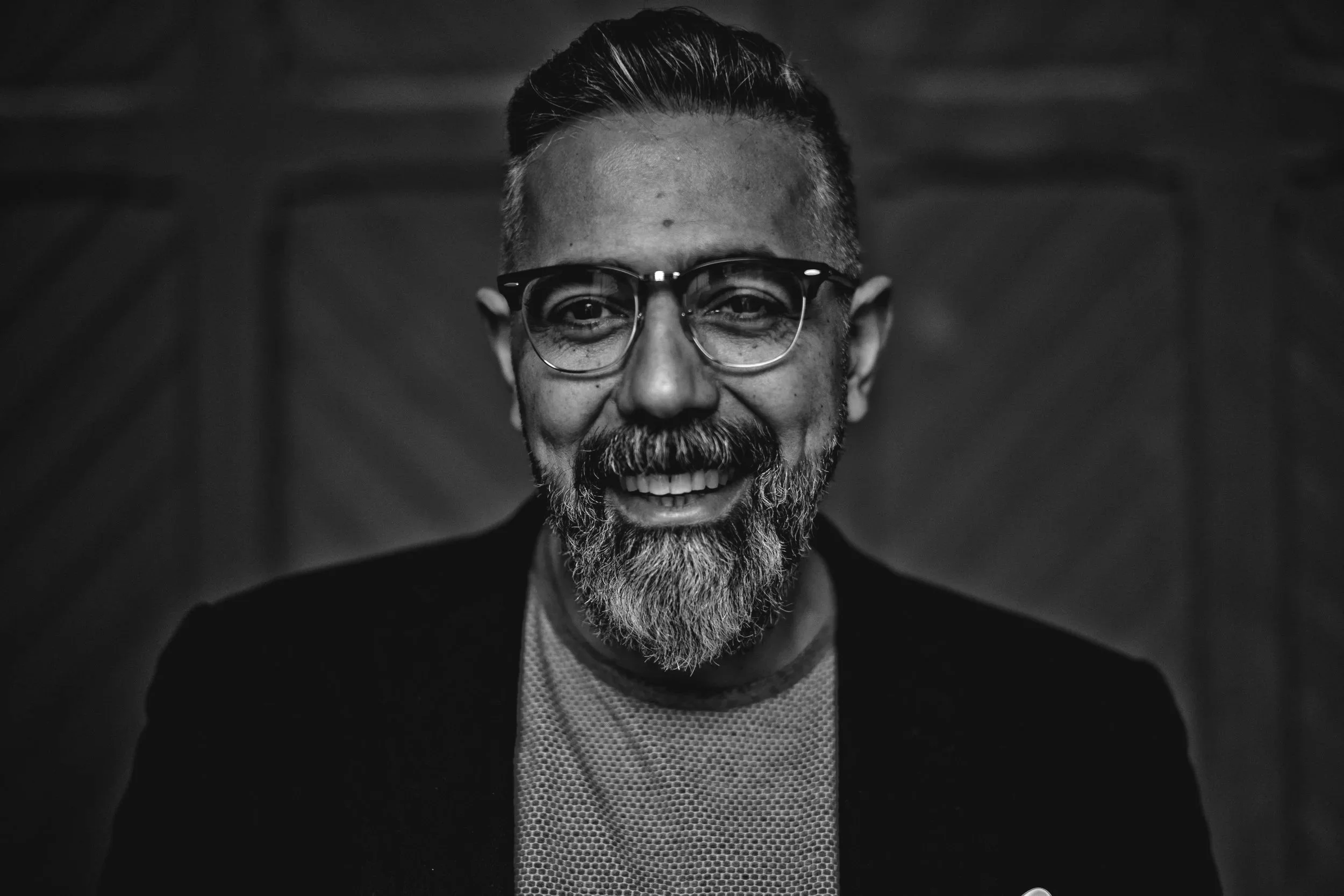 Svartvitt foto av en revisor med glasögon, skägg och varmt leende, mot en trädörr i bakgrunden.