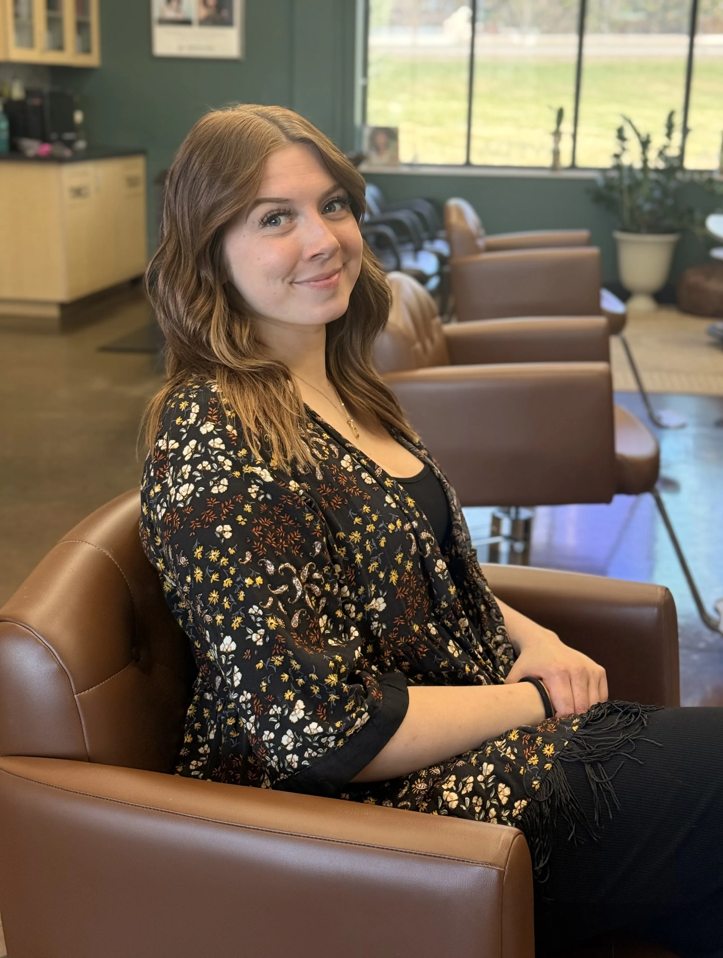 A young woman with wavy brown hair sits on a tan leather chair in a cozy, well-lit room with a large window, black chairs, and plants in the background.