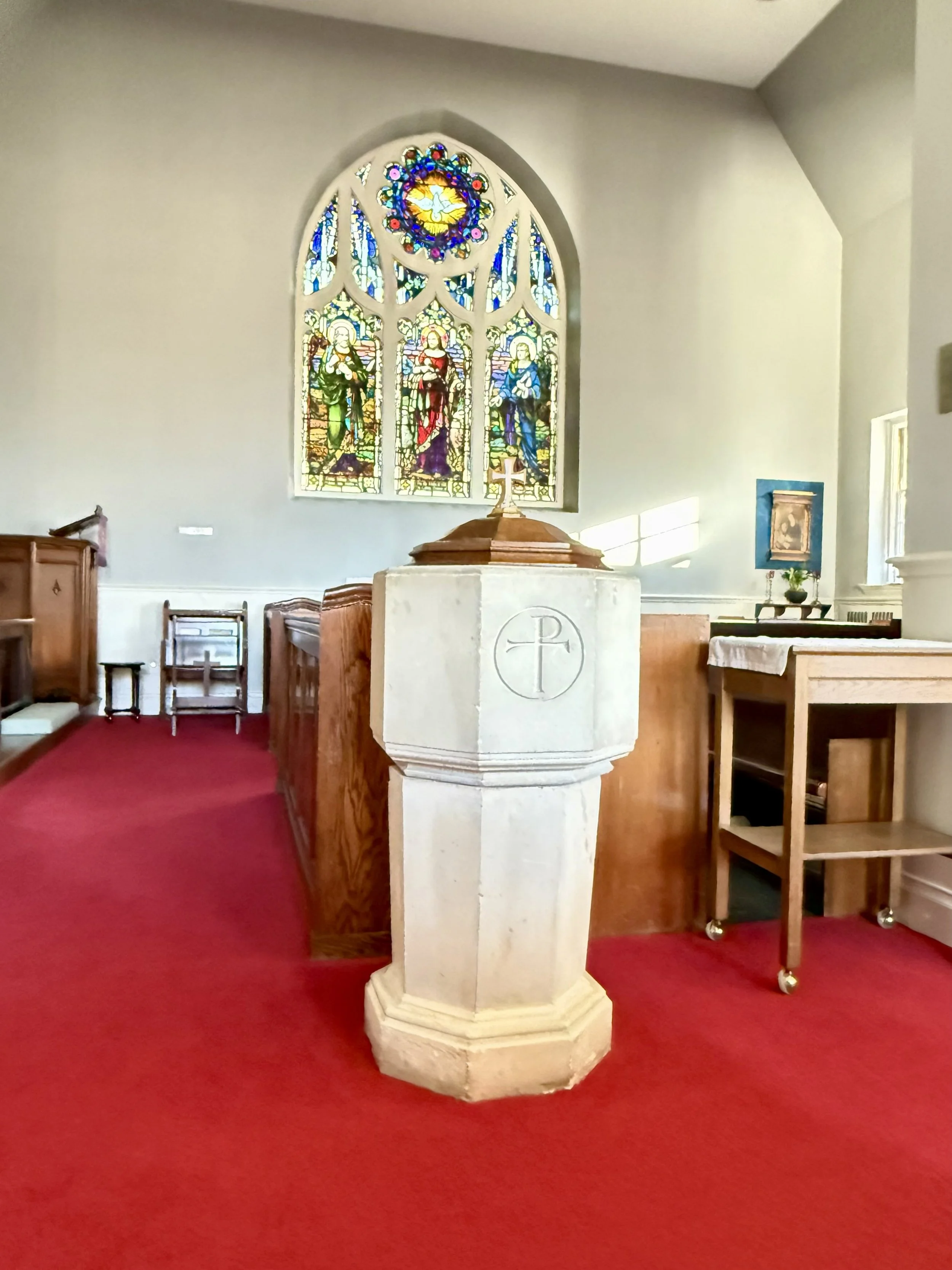 Interior view of a church with a stone baptismal font in the foreground, a stained glass window depicting saints or religious figures in the background, and wooden pews along the sides.