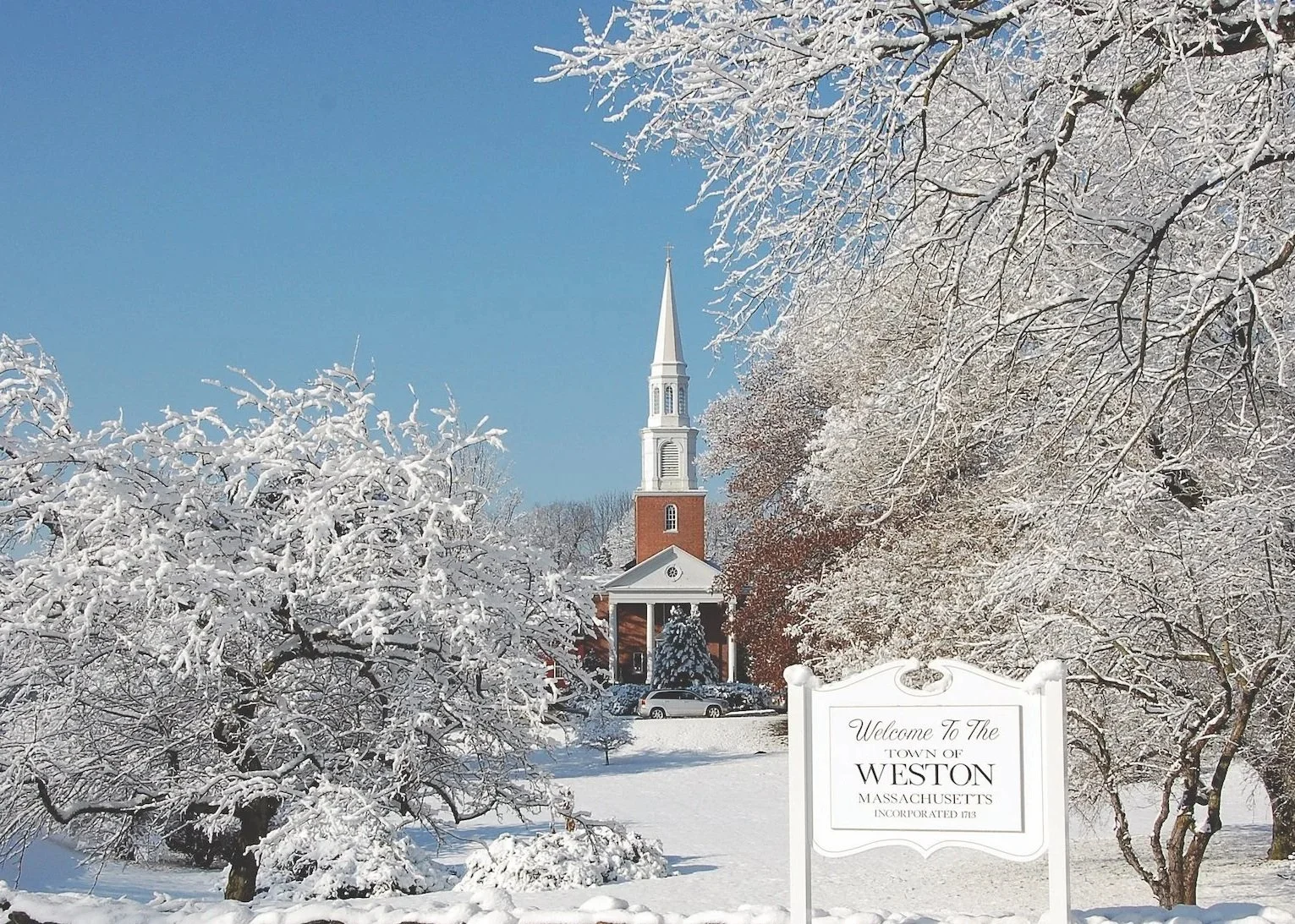 Snow-covered trees and a church with a steeple in a town named Weston, Massachusetts, as indicated by the sign in the foreground. The sky is clear and blue.