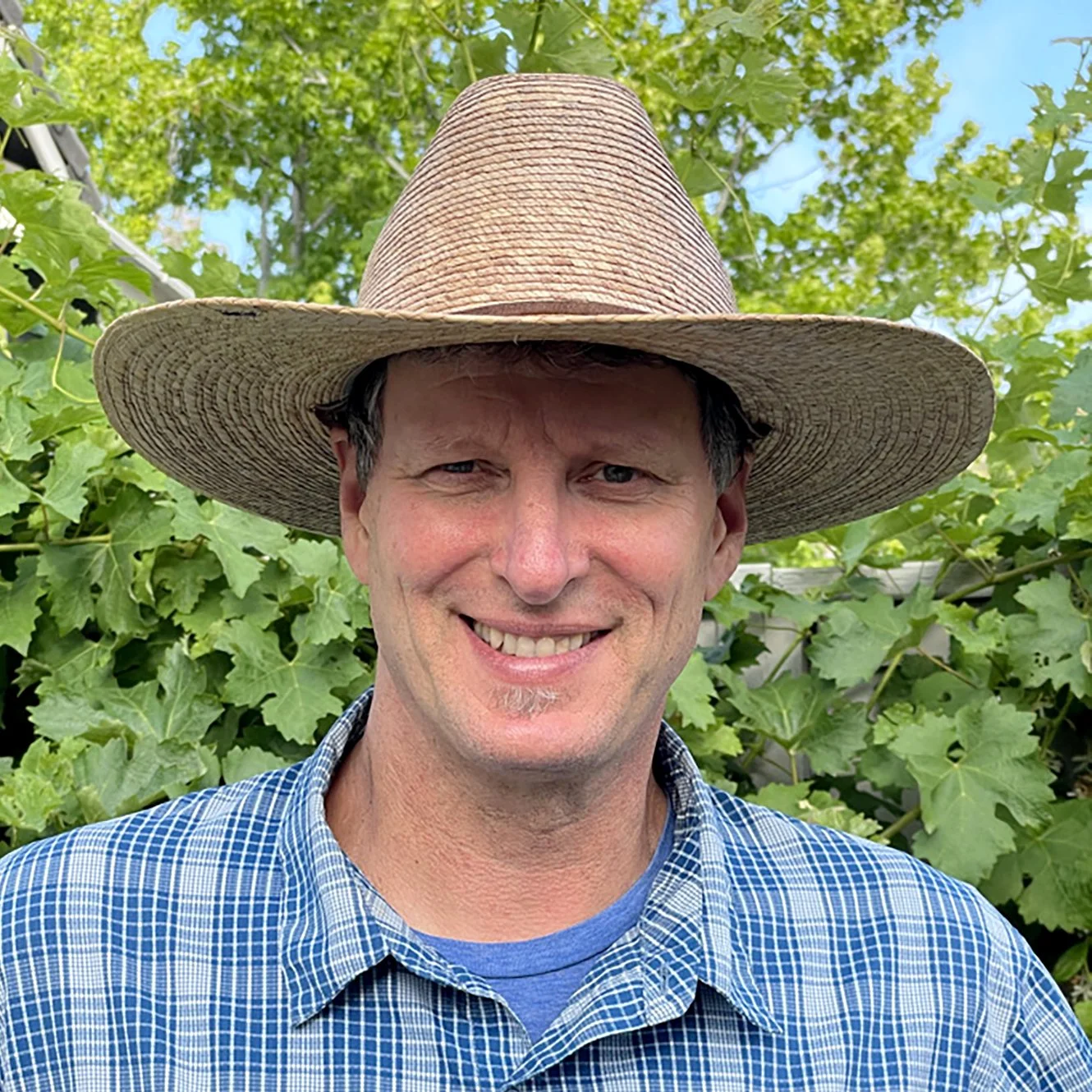 A man smiling outdoors in front of green plants, wearing a large straw hat and a blue checkered shirt.