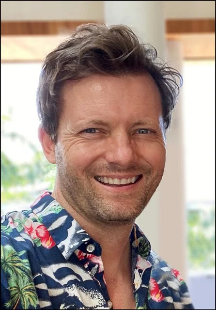 A man with light brown hair and a beard smiling, wearing a colorful Hawaiian shirt, with a blurred indoor background.