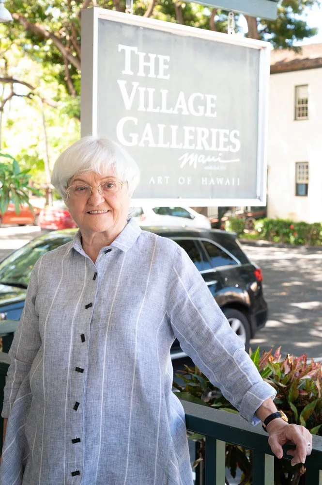 An elderly woman with white hair and glasses wearing a light gray button-up shirt standing outdoors next to a railing with a sign for The Village Galleries in the background. There are trees and parked cars behind her.