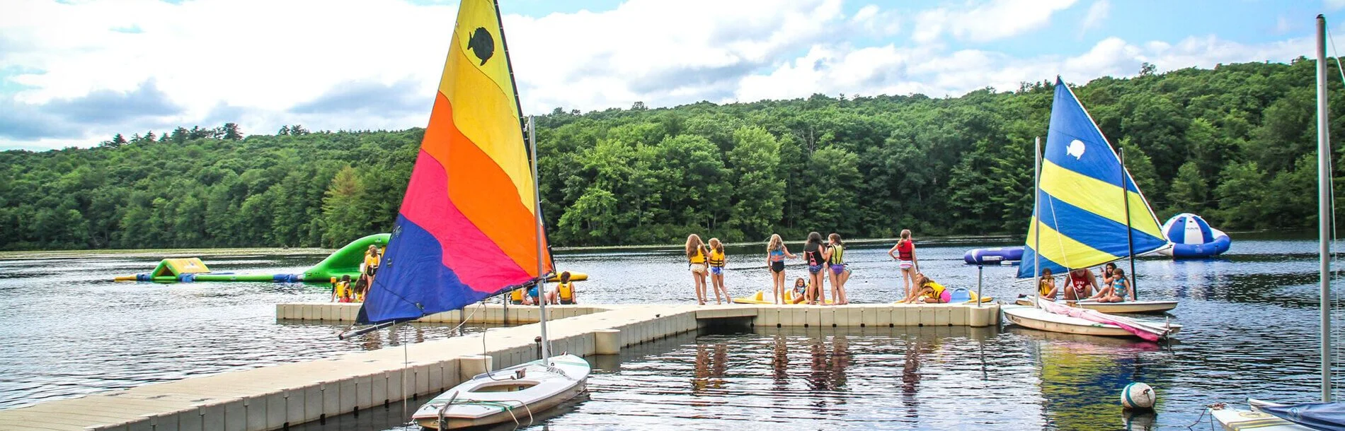 Group of children and teenagers in life jackets on a dock at a lake, preparing for sailing with colorful sailboats, surrounded by lush green trees under a partly cloudy sky.