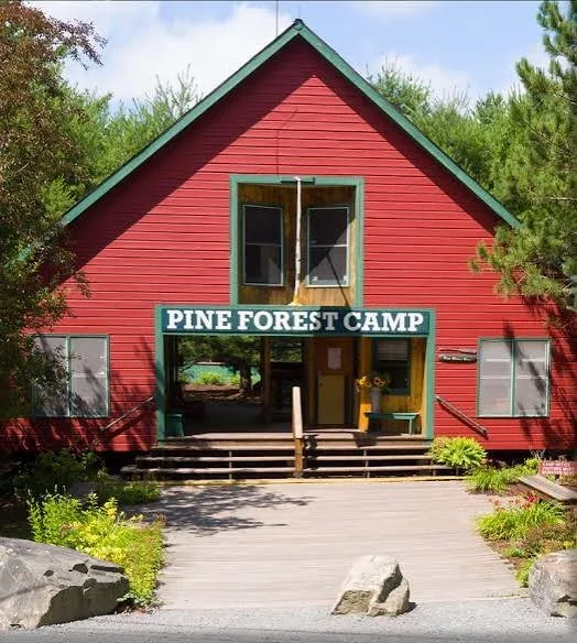 Red wooden building with green trim, labeled Pine Forest Camp, surrounded by trees and shrubs.