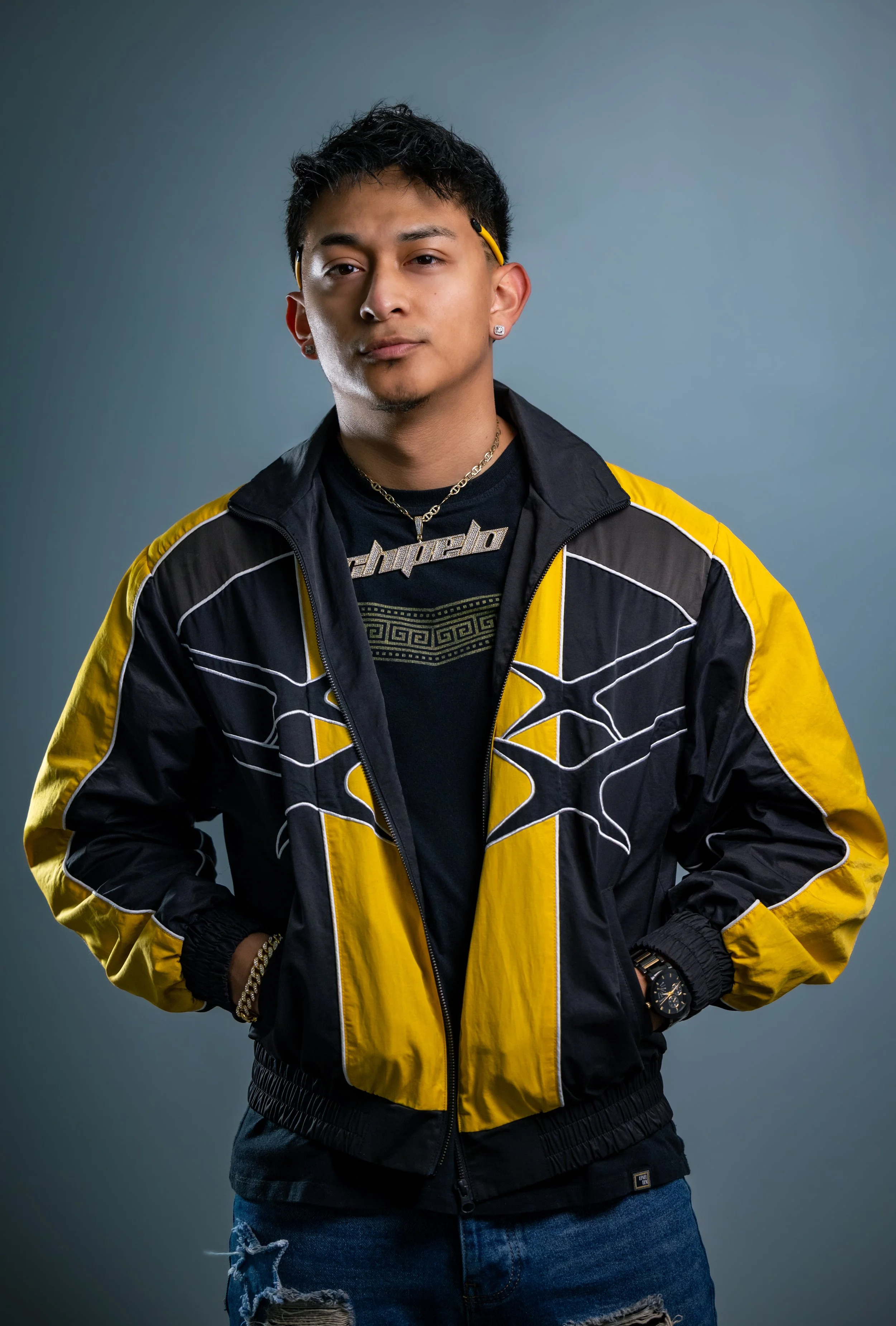 A young man wearing a black and yellow jacket, black t-shirt with a graphic design, and ripped jeans, posing against a plain background.
