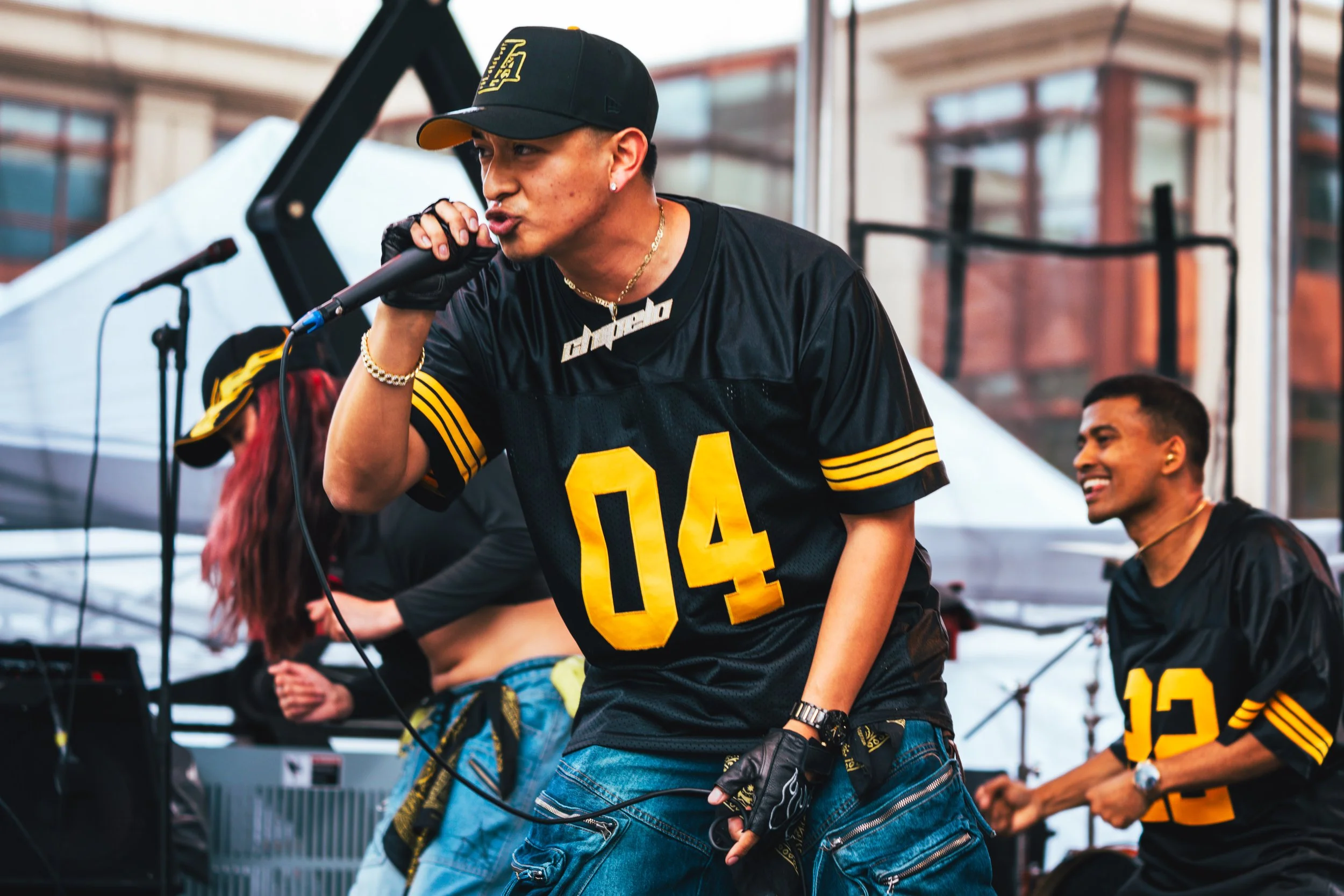 wearing a black and yellow sports jersey with the number 04 and a black cap is singing into a microphone during a music performance. Two other people are seen in the background, also wearing black jerseys with yellow accents, smiling and dancing on an outdoor stage.