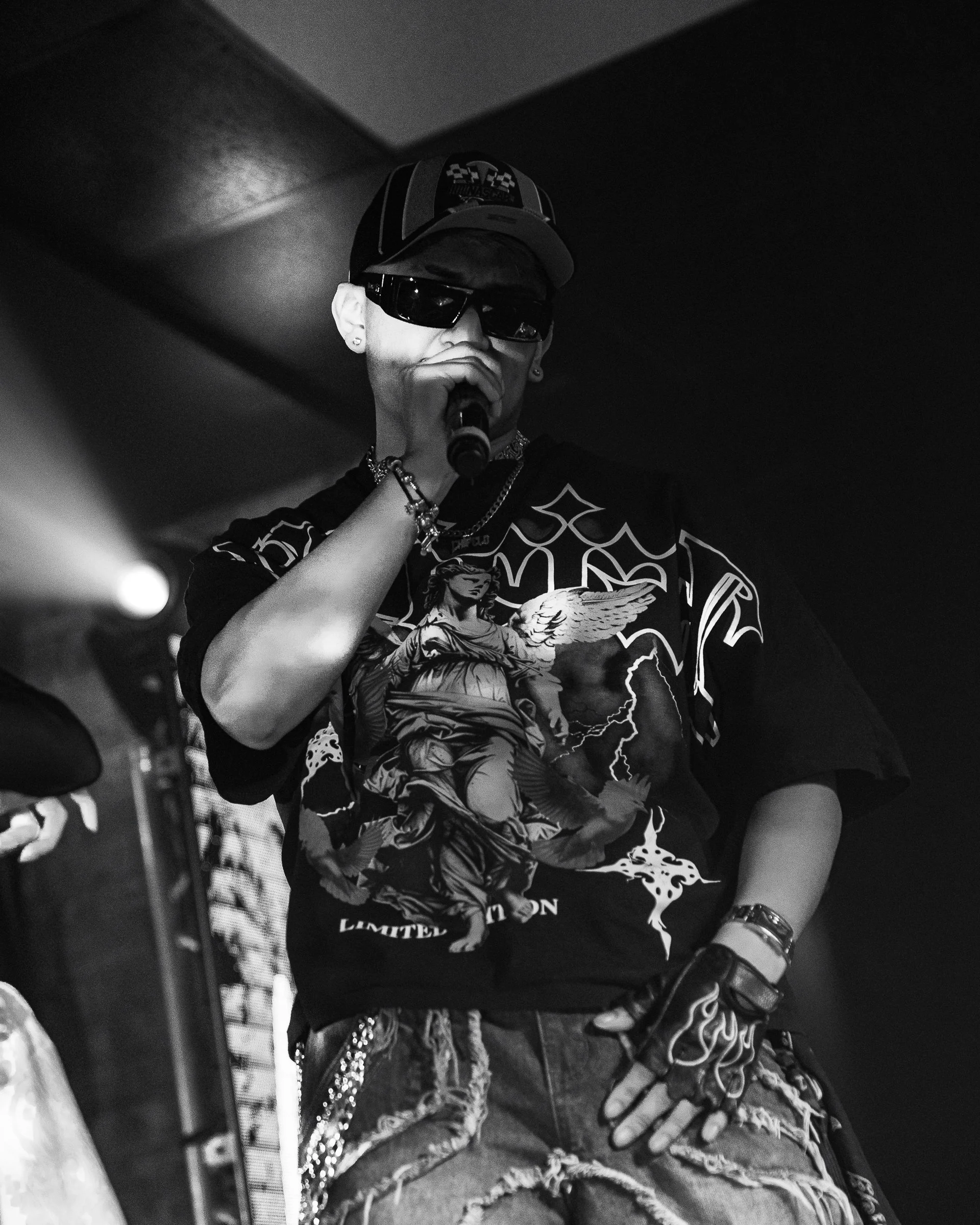 A male performer wearing sunglasses, a cap, a graphic T-shirt, ripped jeans, and accessories, holding a microphone on stage.