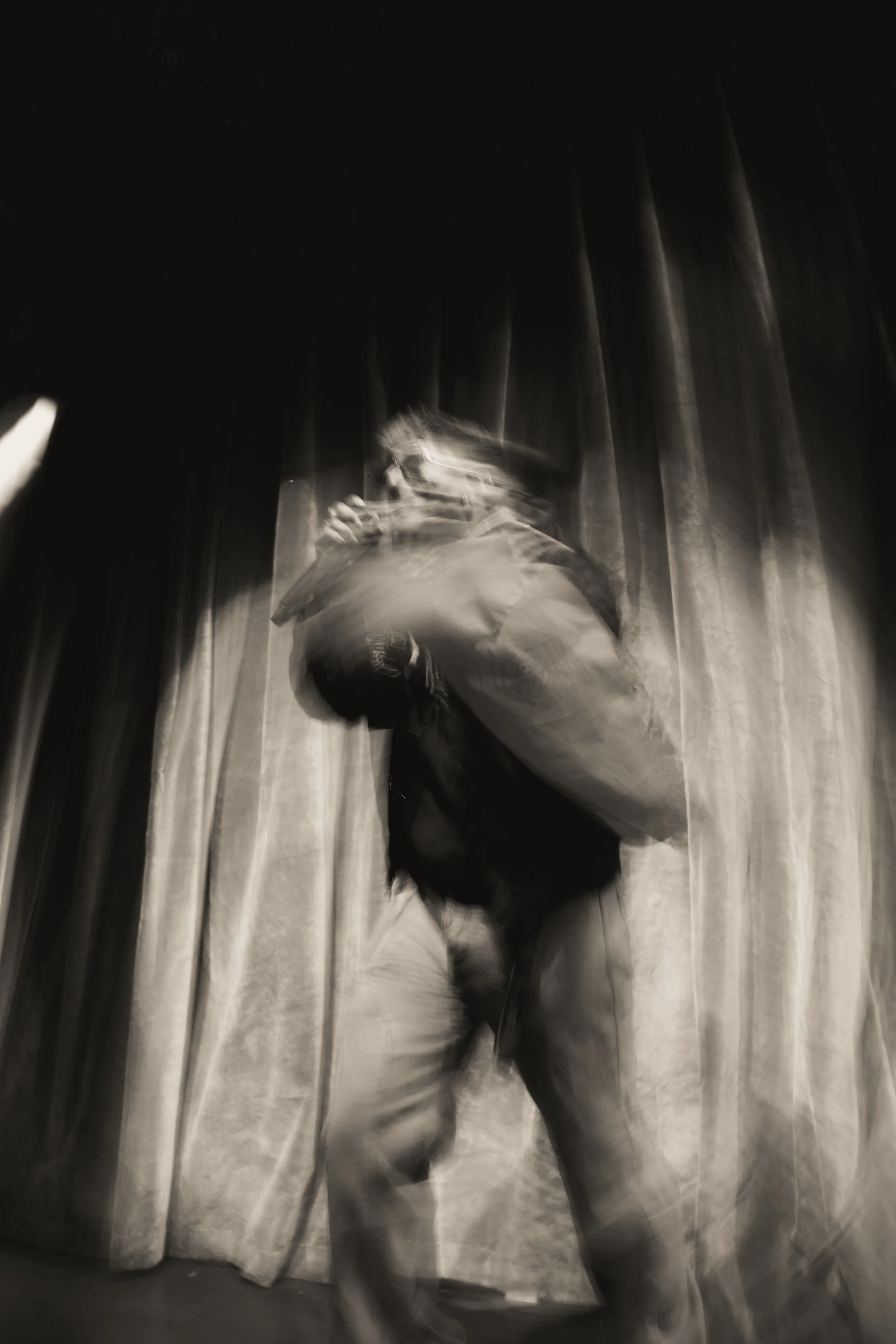 A blurry black-and-white photo of a person on stage holding a microphone and a piece of paper, standing in front of curtains.