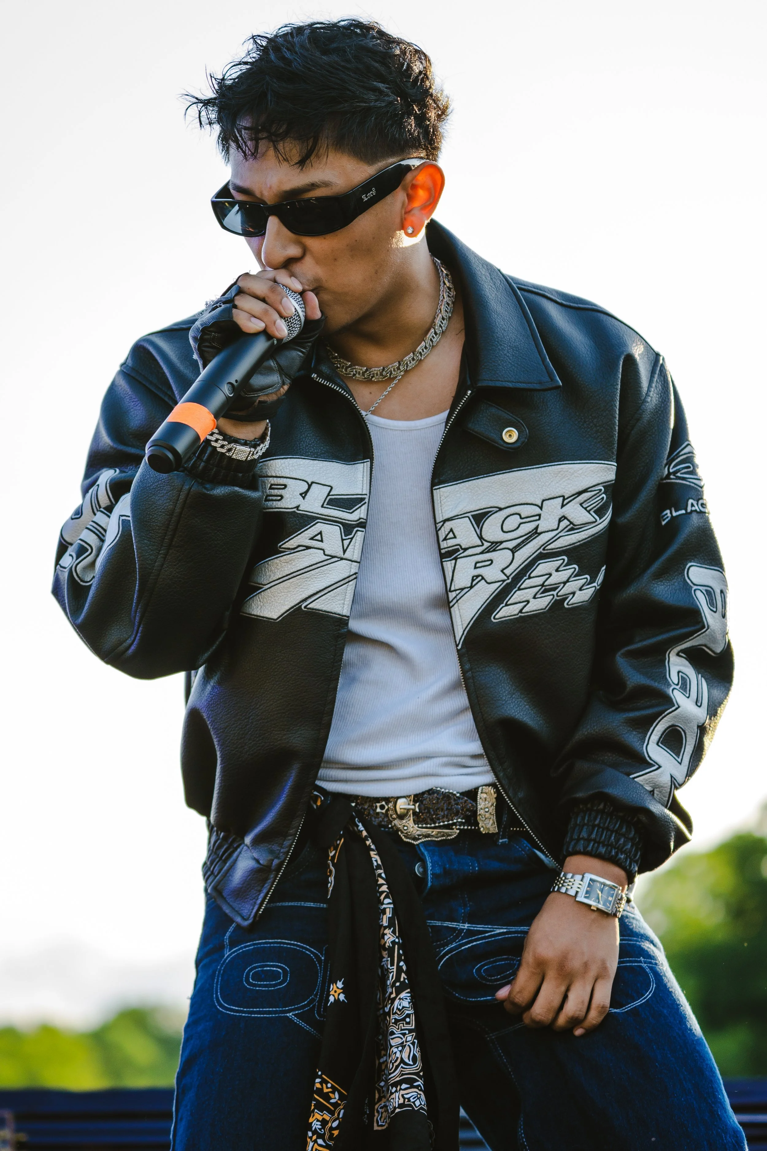 Chipelo singing into a microphone, wearing sunglasses, a black leather jacket, a white t-shirt, jeans, a silver watch, and a patterned scarf around his waist.