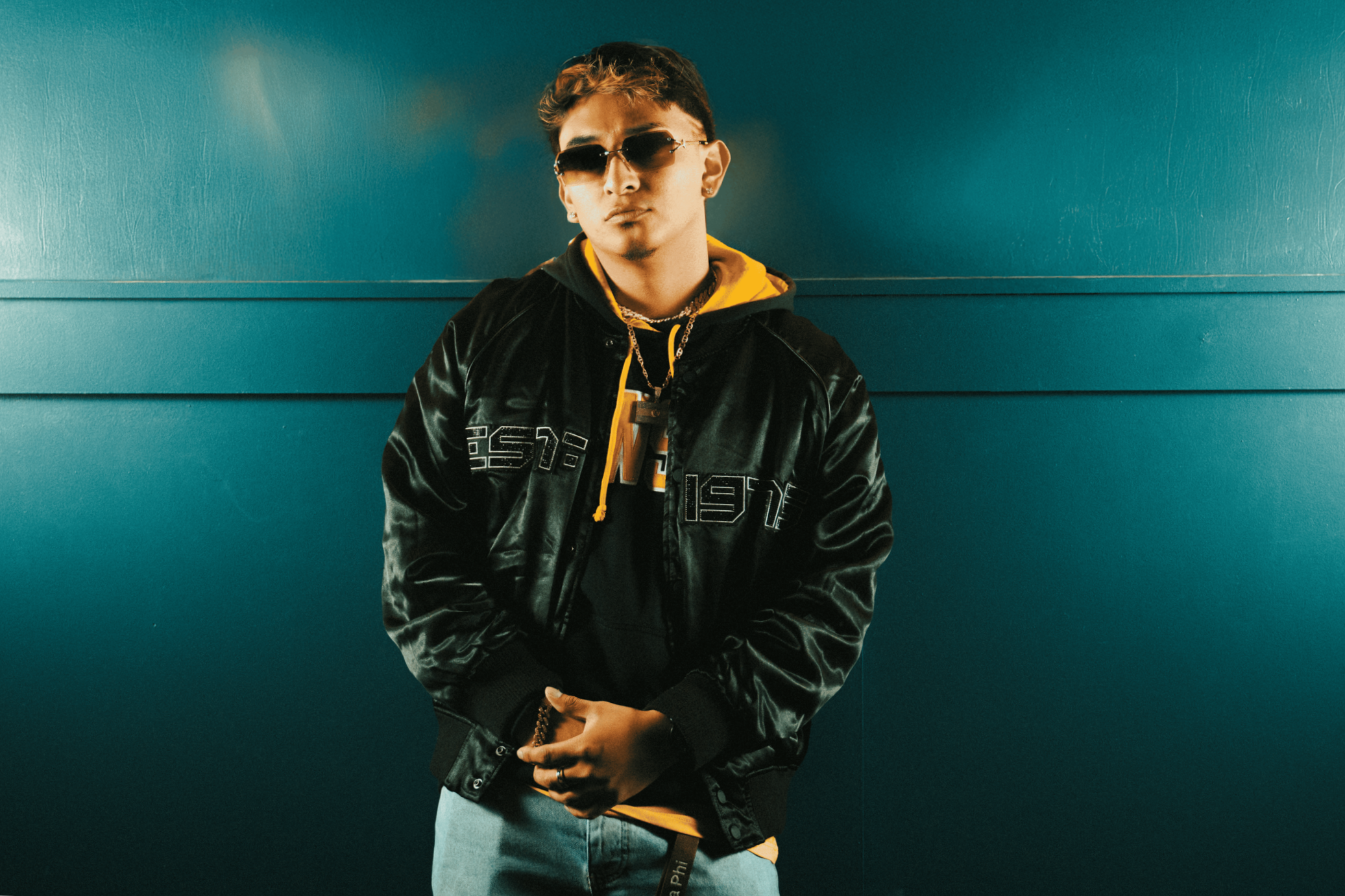 Chipelo wearing sunglasses, a black shiny jacket, and a hoodie with yellow accents, standing against a teal wall.