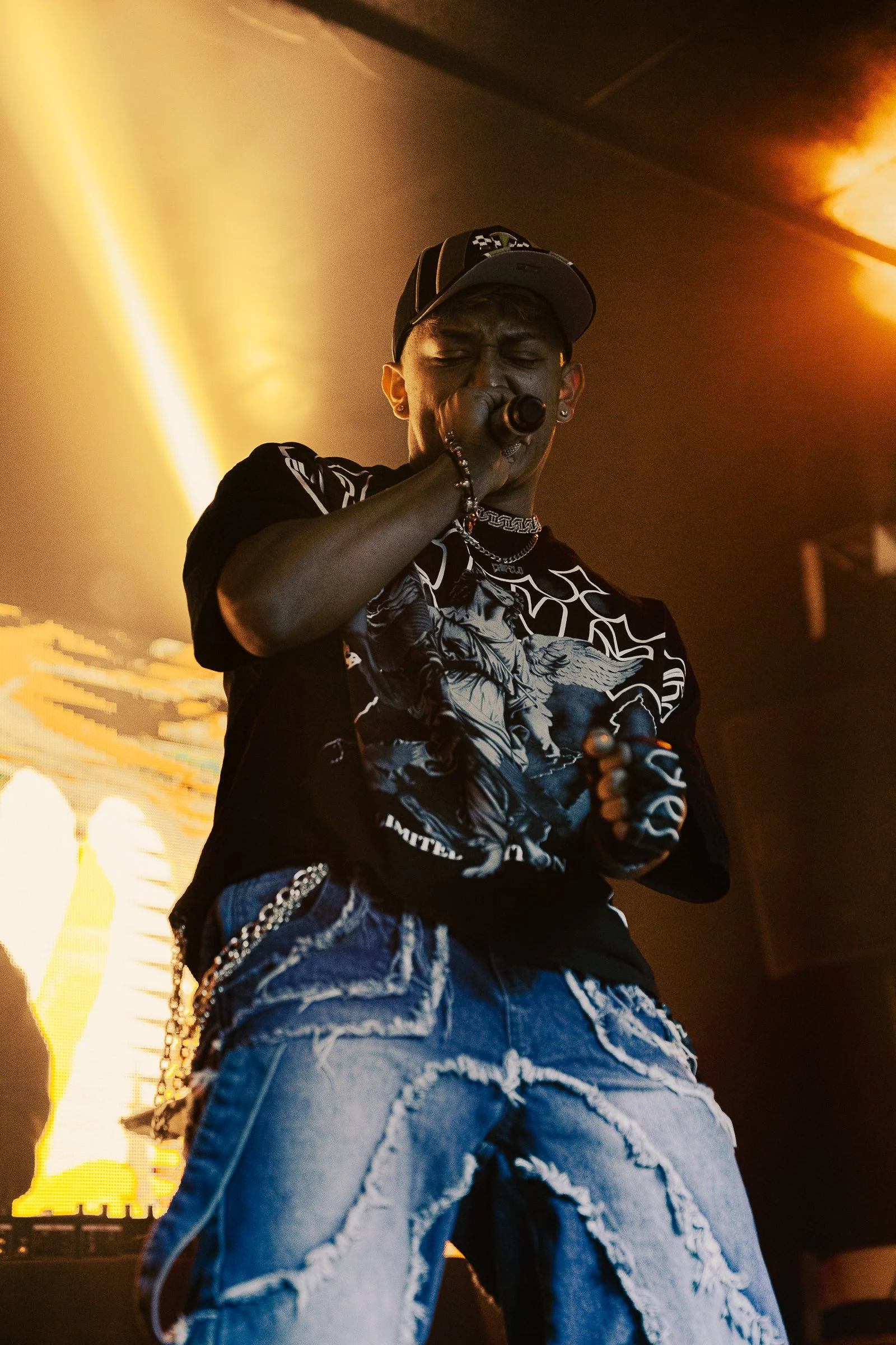 Chipelo singing into a microphone during a live performance, wearing a cap, graphic t-shirt, distressed jeans, and jewelry, with warm stage lighting in the background.