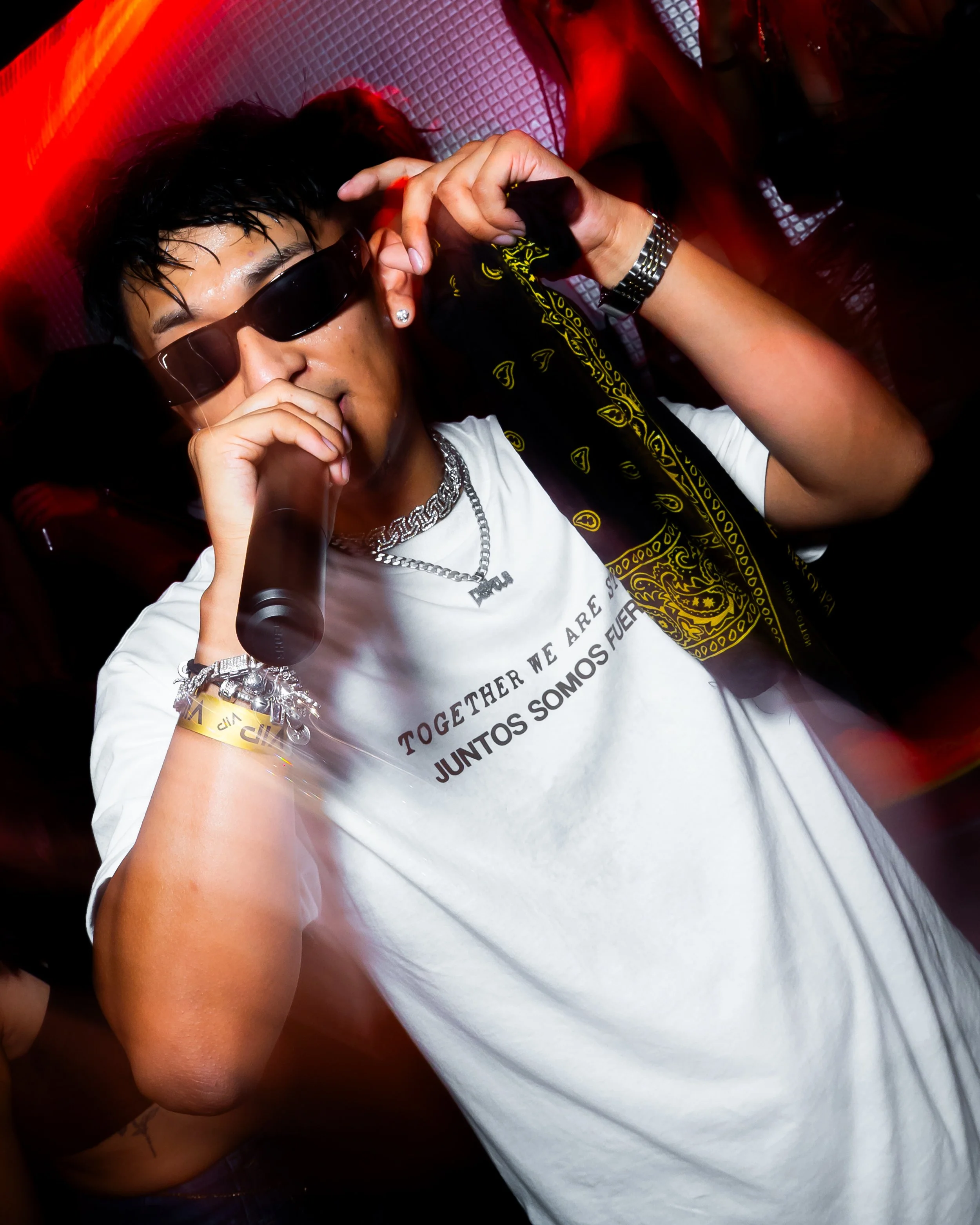 Person wearing sunglasses, holding a microphone, and wearing a white T-shirt with Spanish and English text, in a lively nightclub setting.