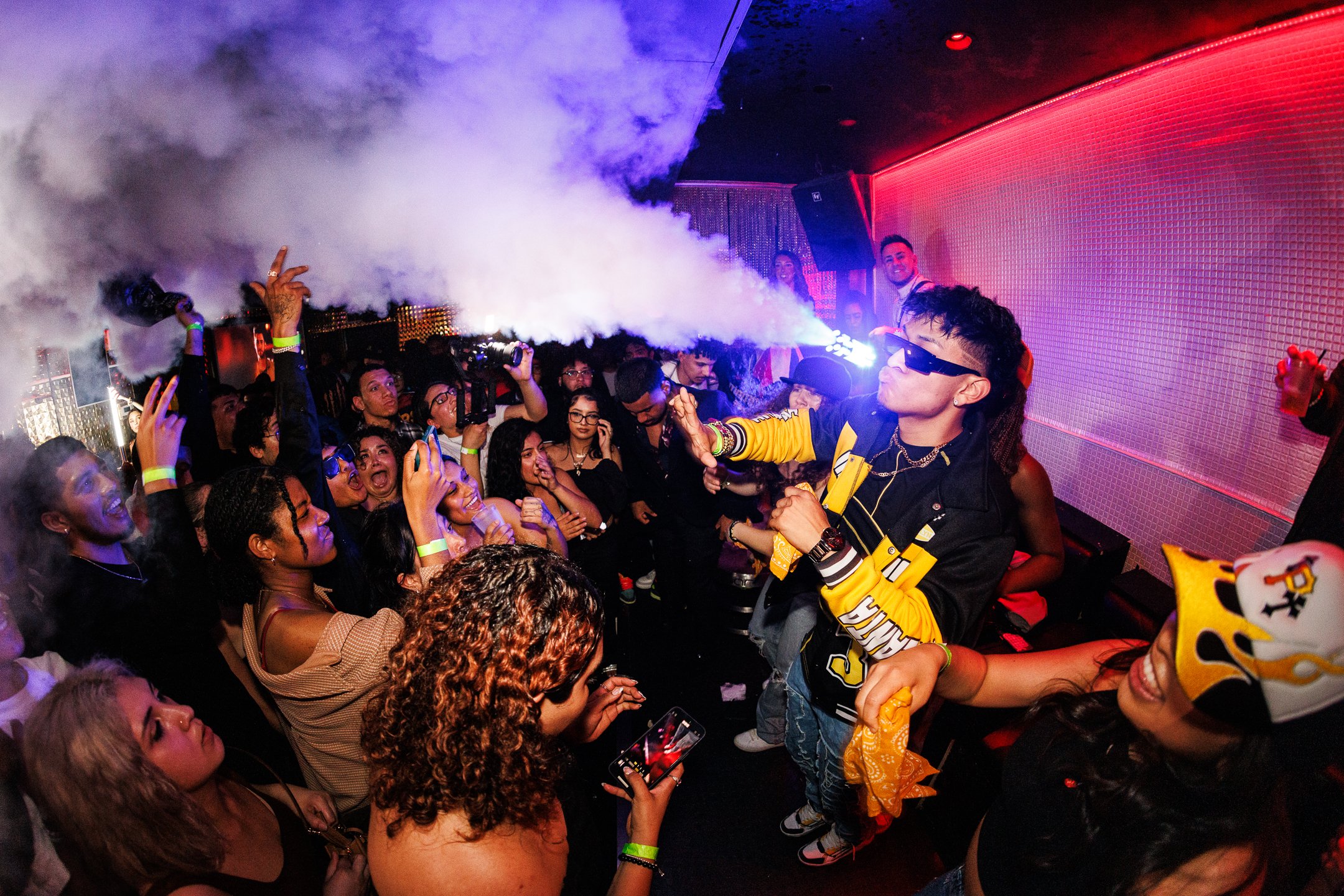 A DJ wearing sunglasses and a yellow and black jacket performs at a nightclub, surrounded by a crowd dancing and enjoying the music, with smoke effects and colorful lighting.
