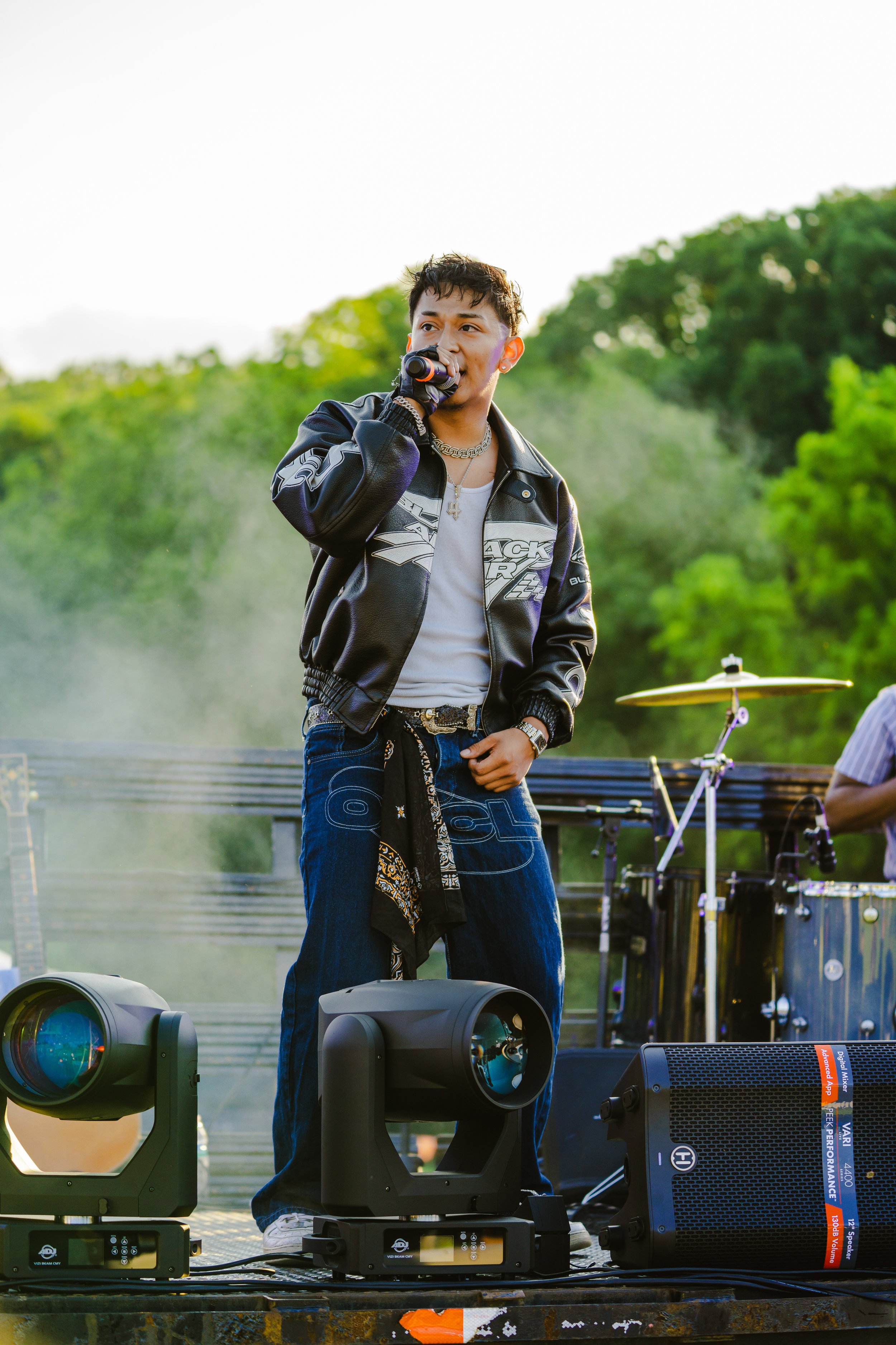 A young male singer performing on an outdoor stage, holding a microphone, surrounded by musical equipment, with greenery in the background.