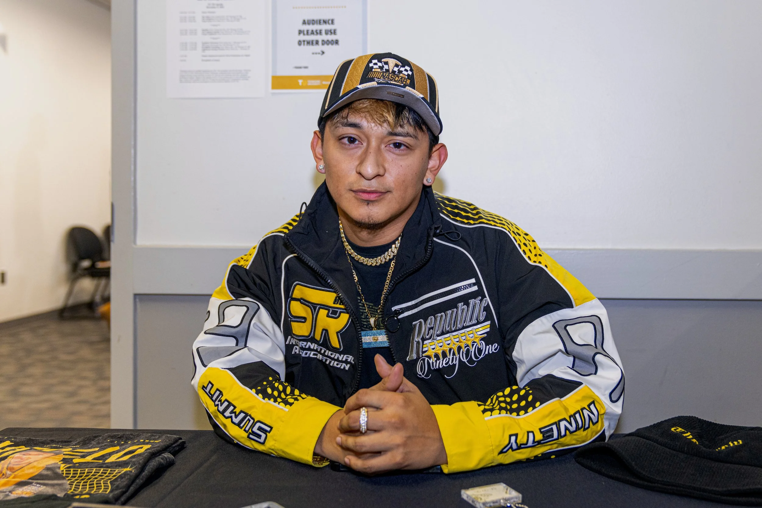 A young man seated at a table wearing a racing jacket and cap with NASCAR logos, with jewelry including rings and necklaces, and two items on the table in front of him.