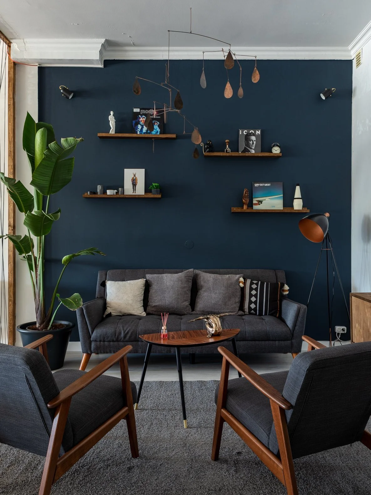 Living room with dark gray sofa, black armchairs with wooden frames, a small wooden coffee table with decorative items, a large green plant, and wall-mounted wooden shelves with various decorative objects, books, and art pieces. The background wall i