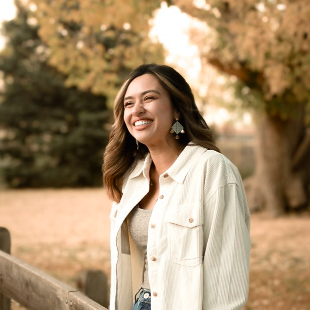 Julieta Vargas headshot (A woman with long, dark, curly hair smiling, wearing a white button down shirt with tan undershirt, standing outdoors in front of a tree.)