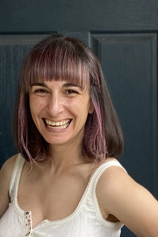 Woman (Irena Y. Shwayder) with shoulder length hair with purple streaks, wearing a white tank top smiling at the camera