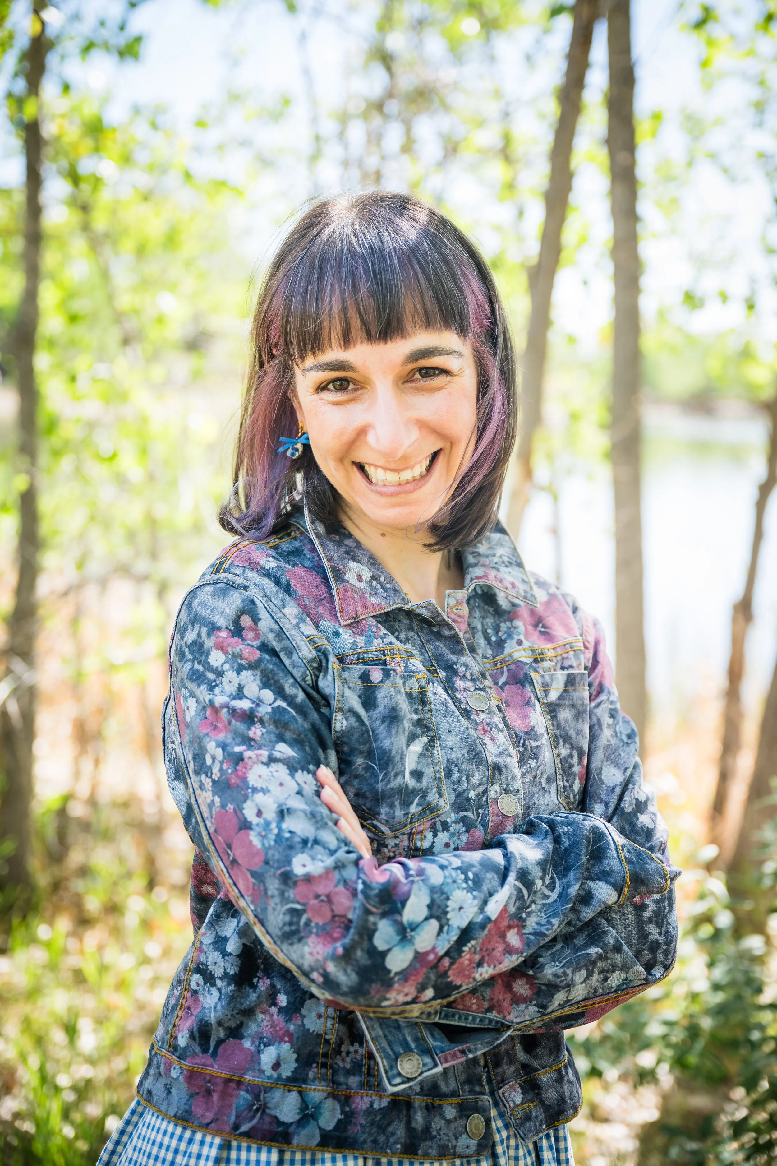 Woman (Irena Y. Shwayder) with shoulder length hair, wearing a flower patterned jacket smiling at the camera while standing in front of trees