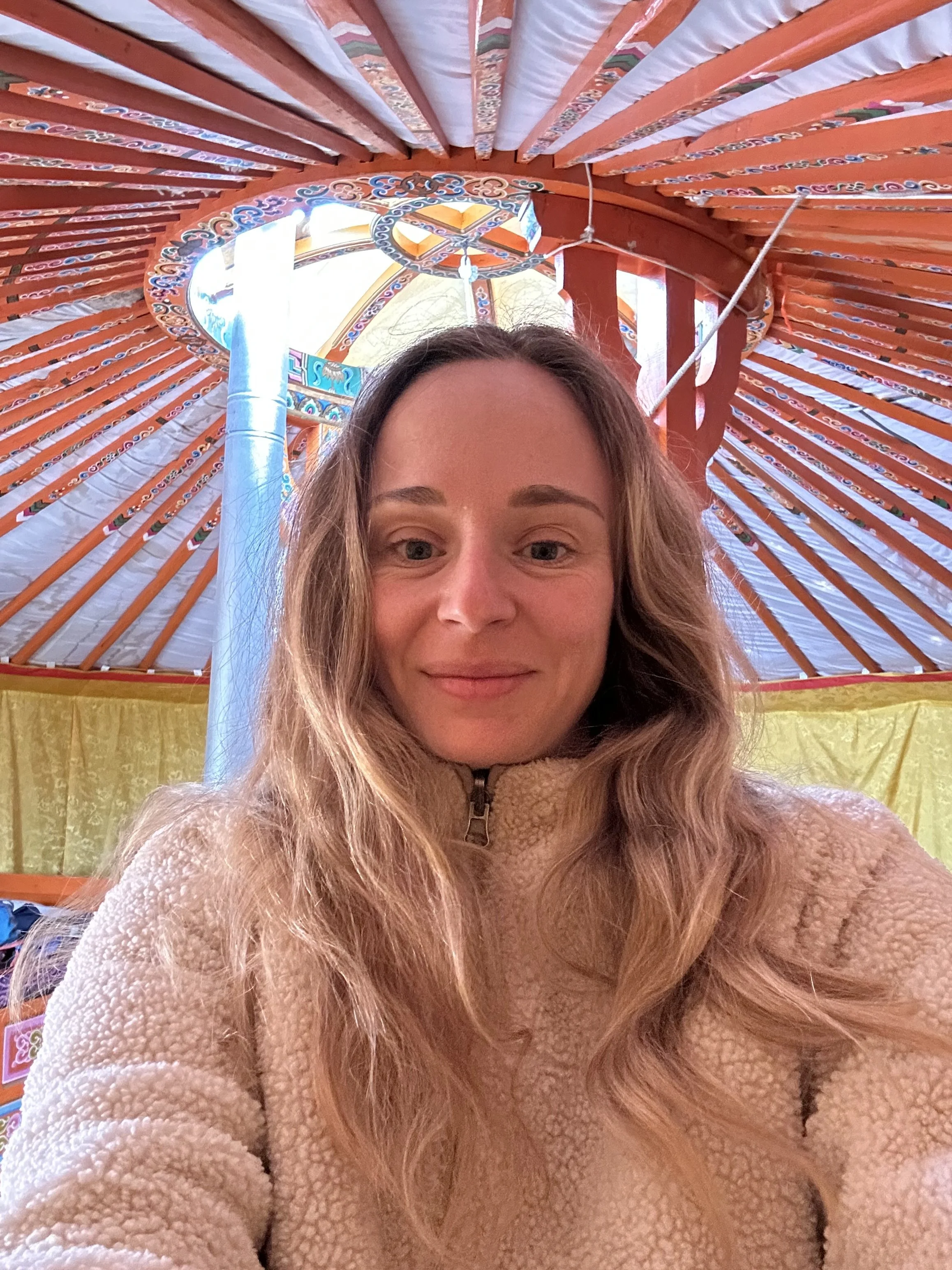 Une femme souriante avec de longs cheveux blonds à l'intérieur d'une structure colorée, ressemblant à une tente ou une grande parapluie, avec un toit richement décoré en bois peint en orange, bleu et blanc.