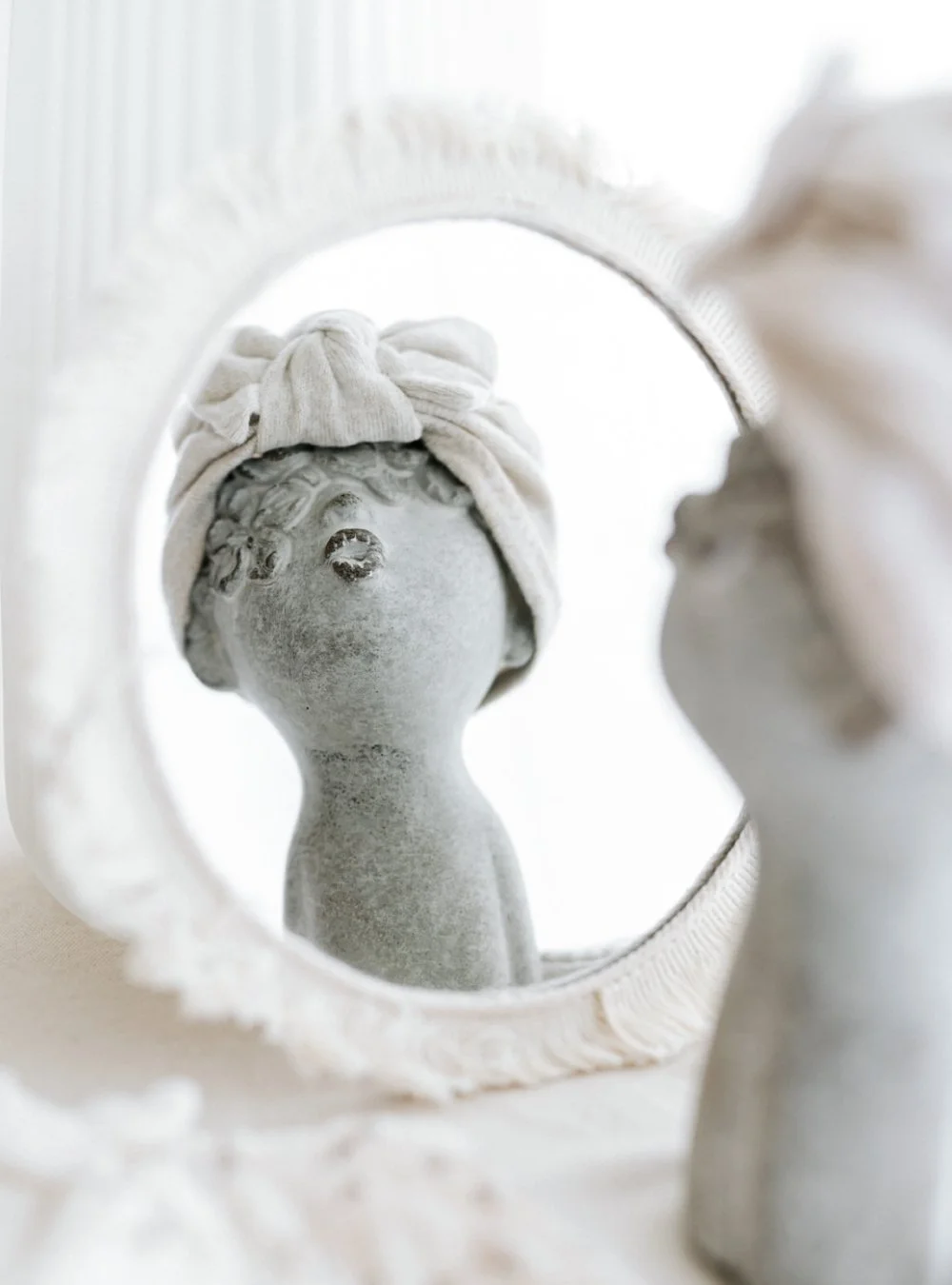 Statue en pierre d'une jeune femme avec un linge blanc enroulé autour de sa tête, vue à travers un miroir ovale.